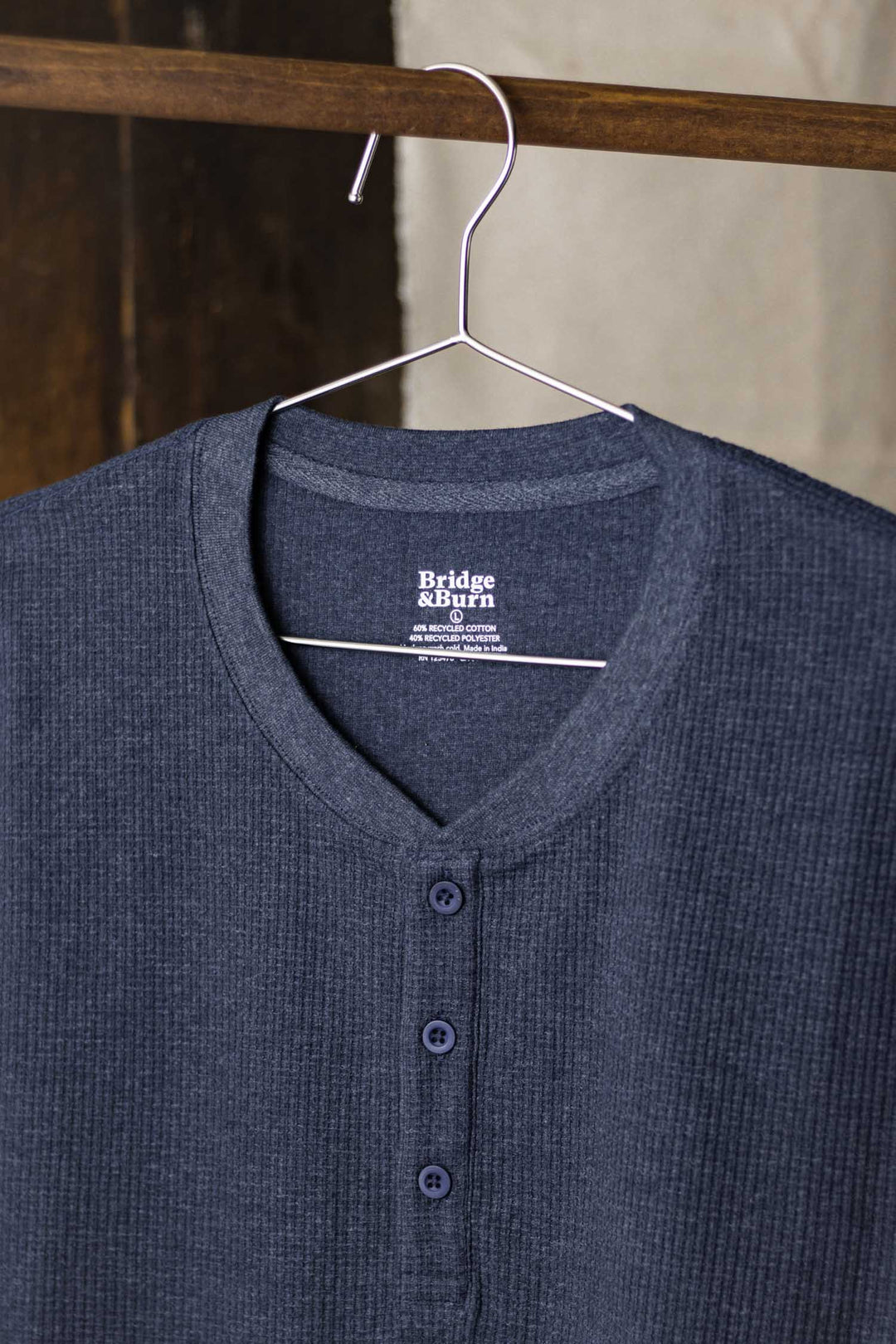 The Women's Recycled Waffle Henley in Sapphire Melange by Bridge & Burn, featuring a three-button placket and thermal waffle texture, hangs on a metal hanger against a blurred wooden and light-toned background.