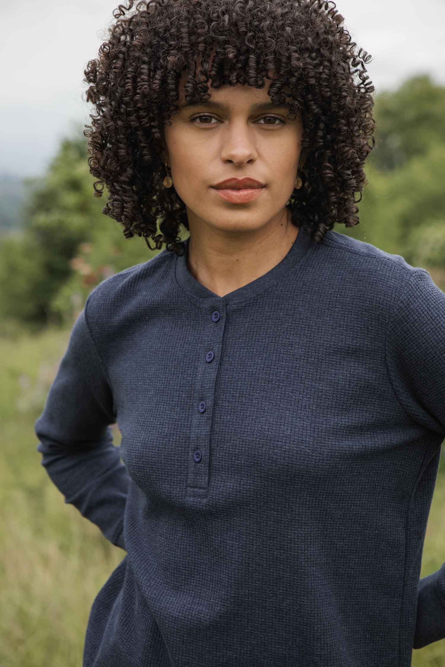 A woman with curly dark hair wears the Bridge & Burn Women's Recycled Waffle Henley in Sapphire Melange, standing in a grassy field with a neutral expression. Trees and greenery create a blurred background outdoors.