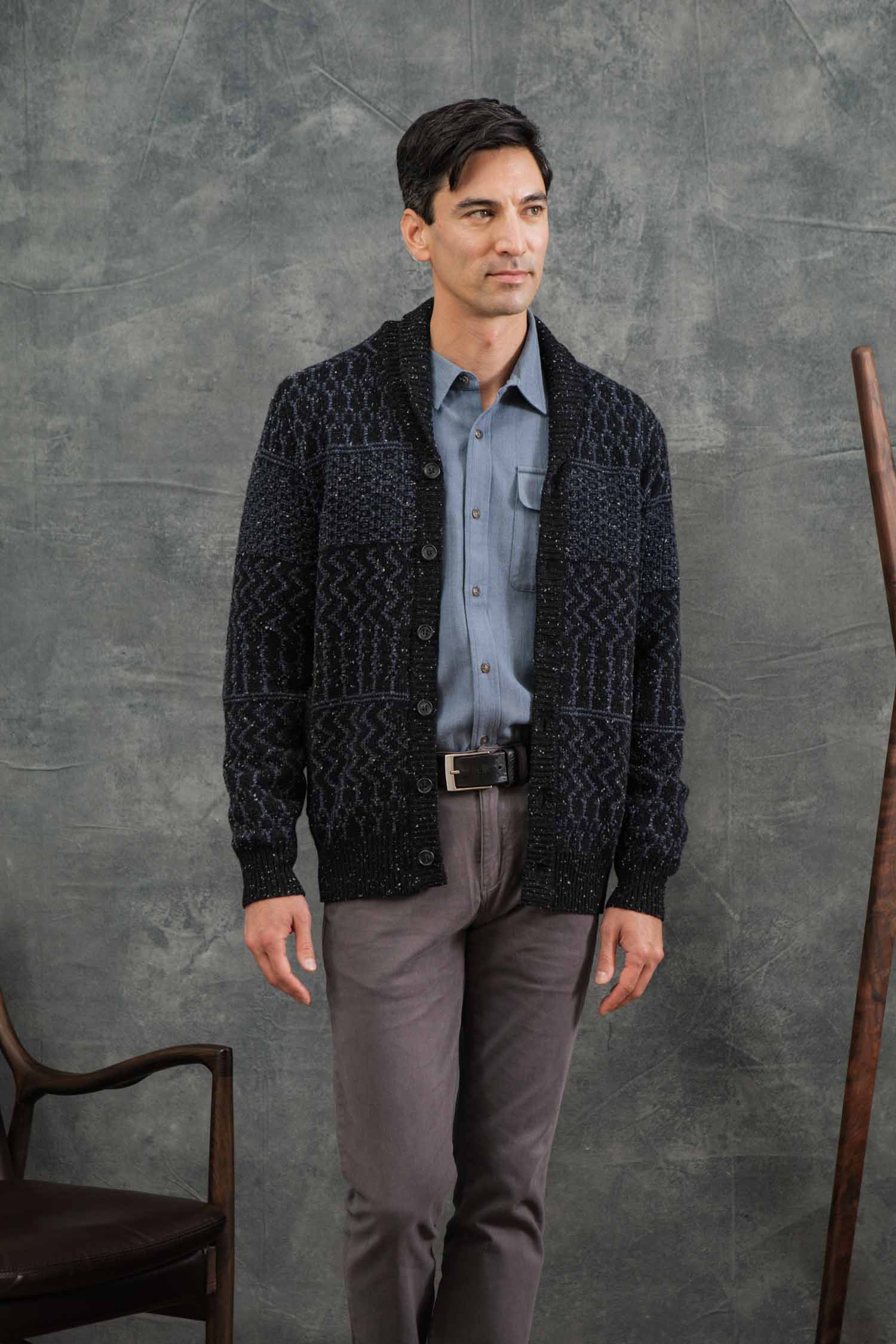 A man with short dark hair stands indoors near a chair and wooden rod, wearing the Bridge & Burn Anders Cardigan in Midnight Tweed (RWS certified wool) over a blue shirt and gray pants, set against a textured gray background.