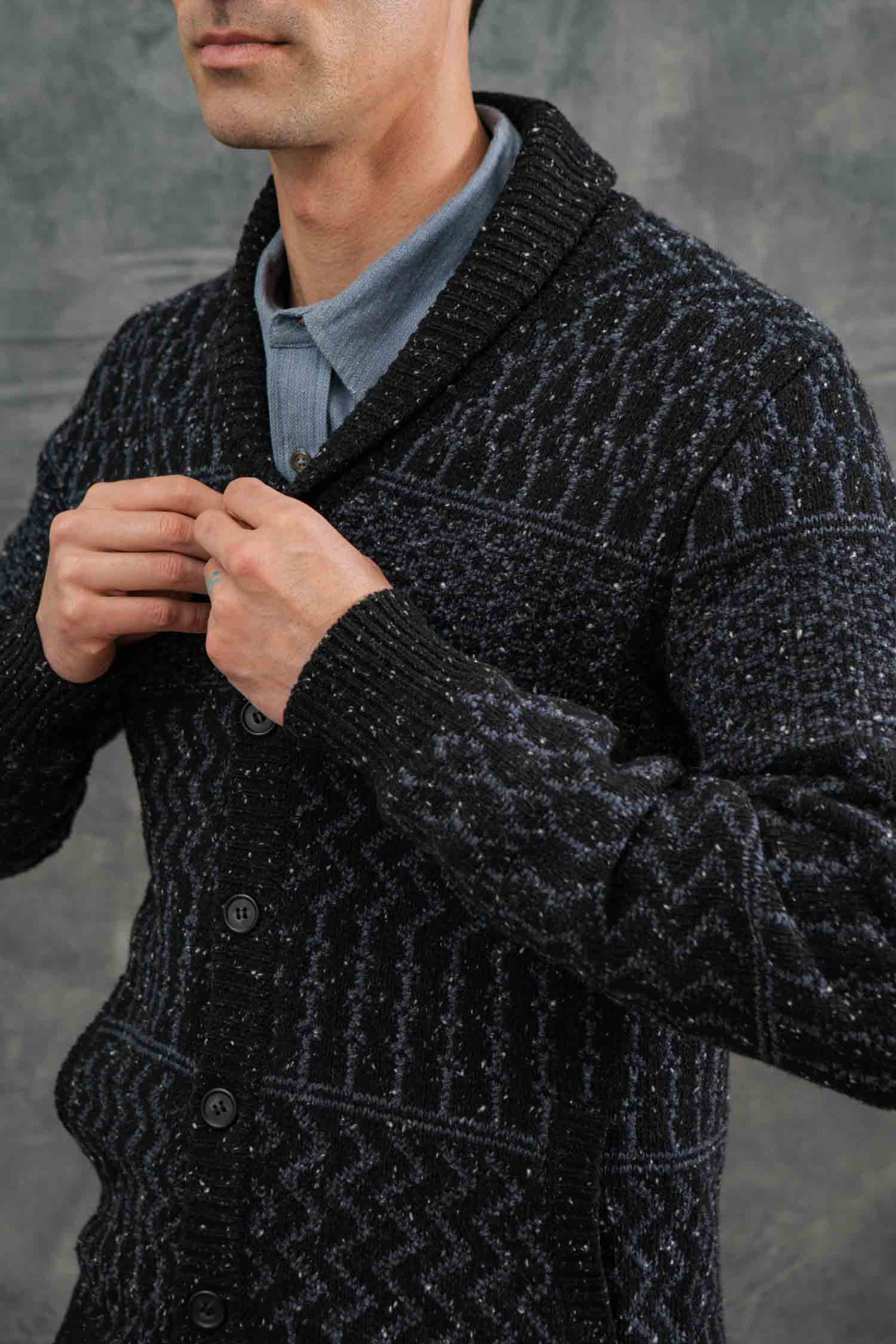 A man, with only the lower half of his face visible, buttons up the Bridge & Burn Anders Cardigan in Midnight Tweed over a light blue collared shirt against a neutral gray background.