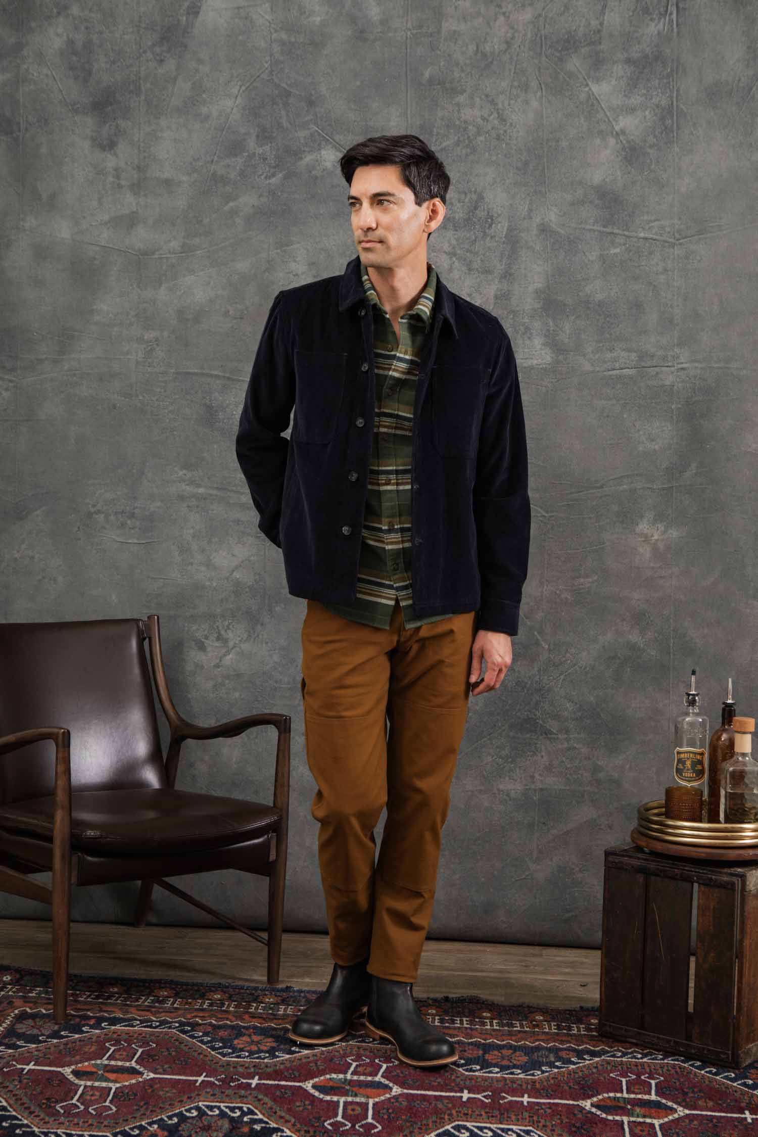 A man stands indoors on a patterned rug, wearing the Bridge & Burn Banks Shirt Jacket in Midnight Blue Corduroy over a green plaid shirt, tan pants, and black boots. He is beside a brown chair and small wooden table with bottles and glasses, against a gray wall.