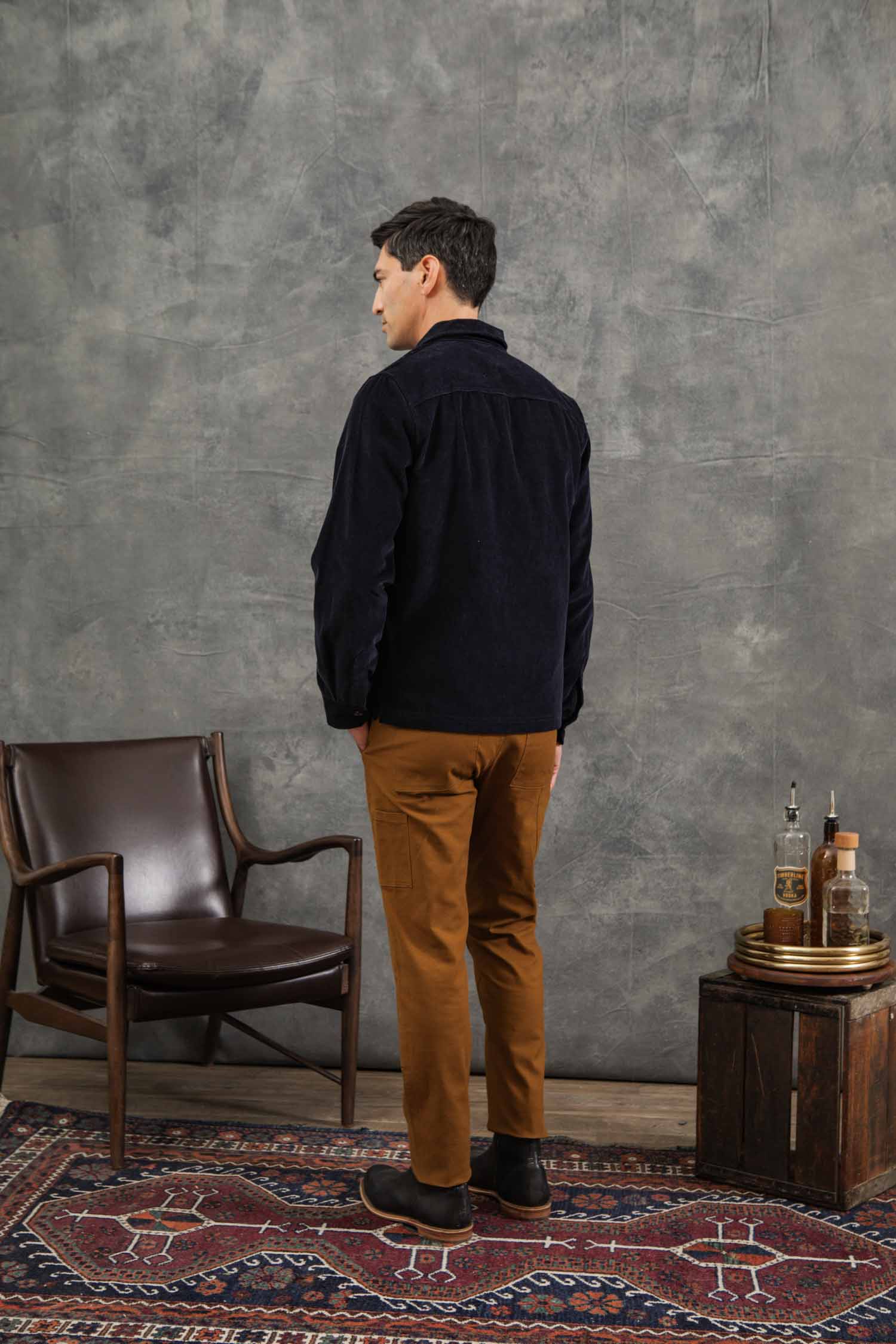 A man with short dark hair stands facing away, wearing the Bridge & Burn Banks Shirt Jacket in midnight blue corduroy and brown pants. He is beside a brown leather chair and small table with bottles, on a rug against a textured gray wall.