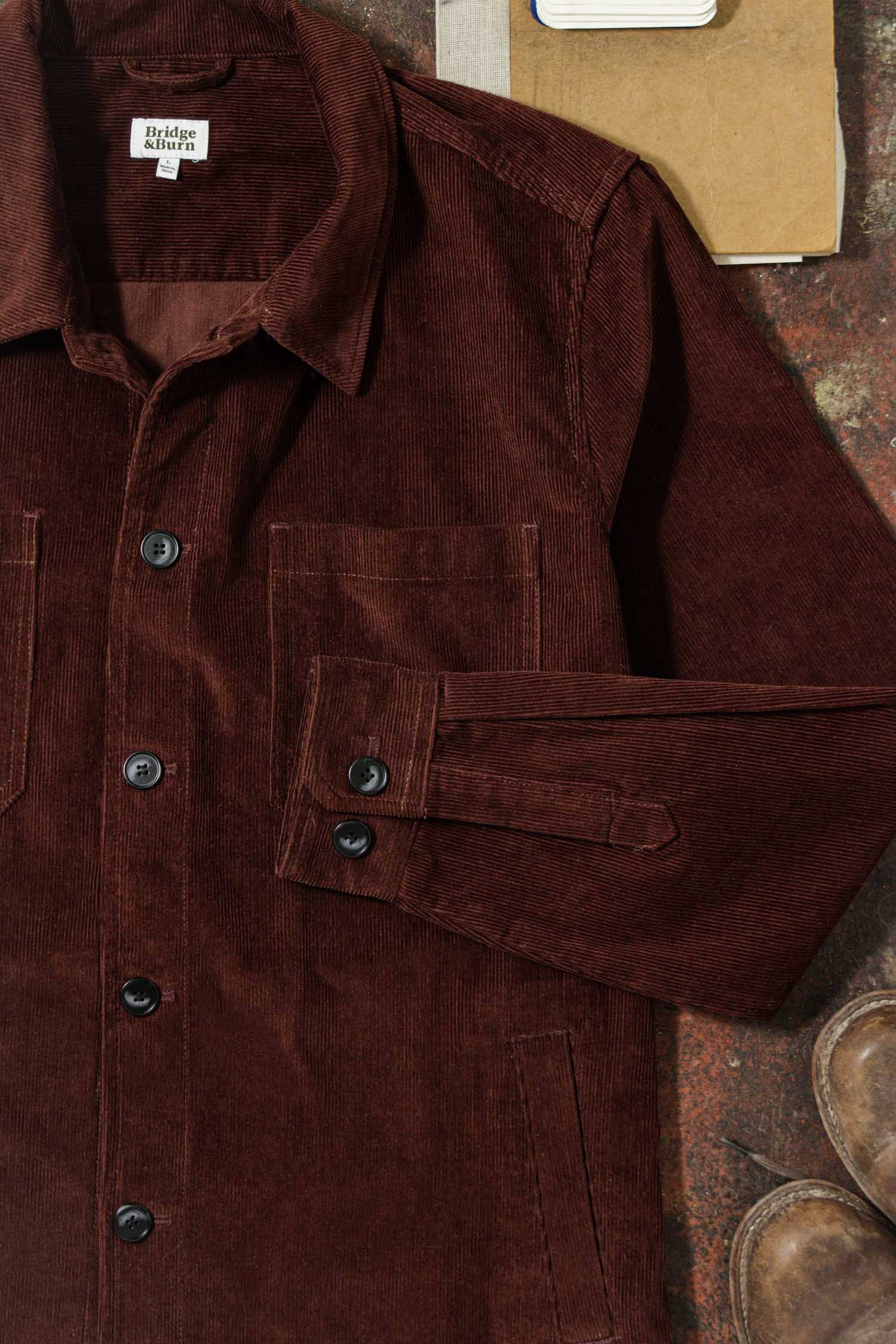 The Bridge & Burn Banks Shirt Jacket in Redwood Corduroy, featuring black buttons, a collared neckline, two front pockets, and a folded left sleeve, is shown laid flat with part of a boot and notebook for a workwear-inspired look.