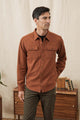 A man with short dark hair wears the Bridge & Burn Bedford Shirt in Terracotta Melange and olive green pants, standing indoors before a light-colored curtain and a wooden table topped with stacked books.