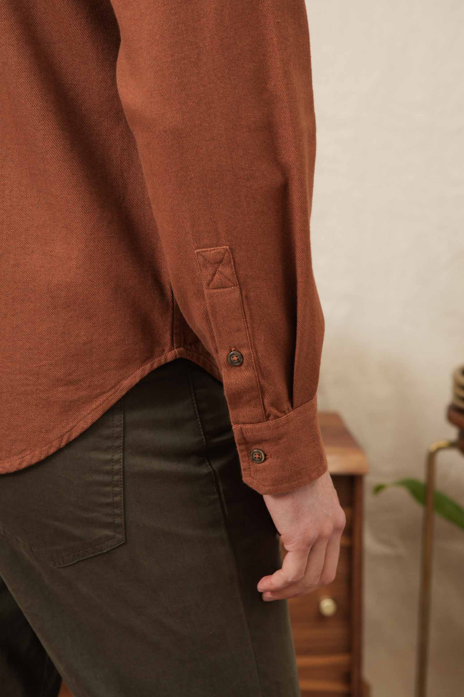 A close-up of a person’s arm and hand wearing the Bridge & Burn Bedford Shirt in Terracotta Melange with dark olive-green pants. A small wooden table and a plant appear in the background.