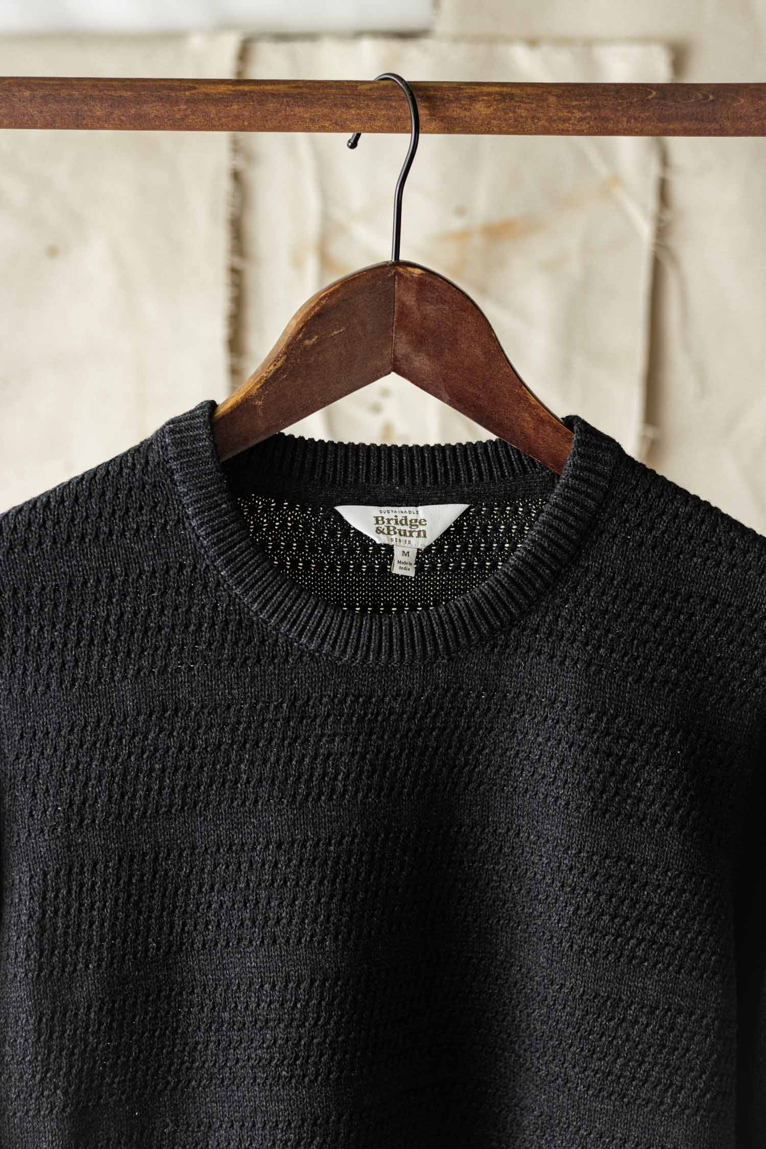 The Bridge & Burn Calder Sweater in Midnight, featuring a textured knit design, hangs on a wooden hanger from a rod against a neutral fabric backdrop.
