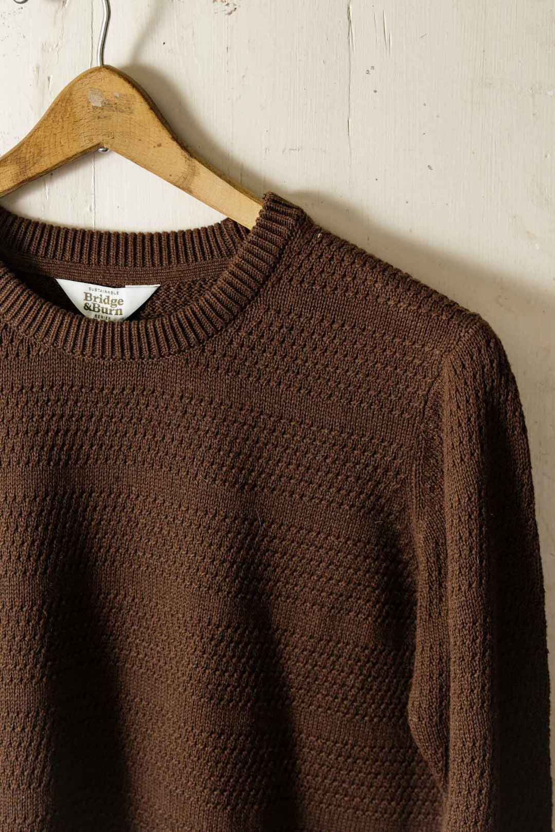 The Calder Sweater in Walnut by Bridge & Burn hangs on a wooden hanger against a light, slightly worn wooden wall. This eco-friendly sweater features a ribbed neckline and subtle textured pattern.