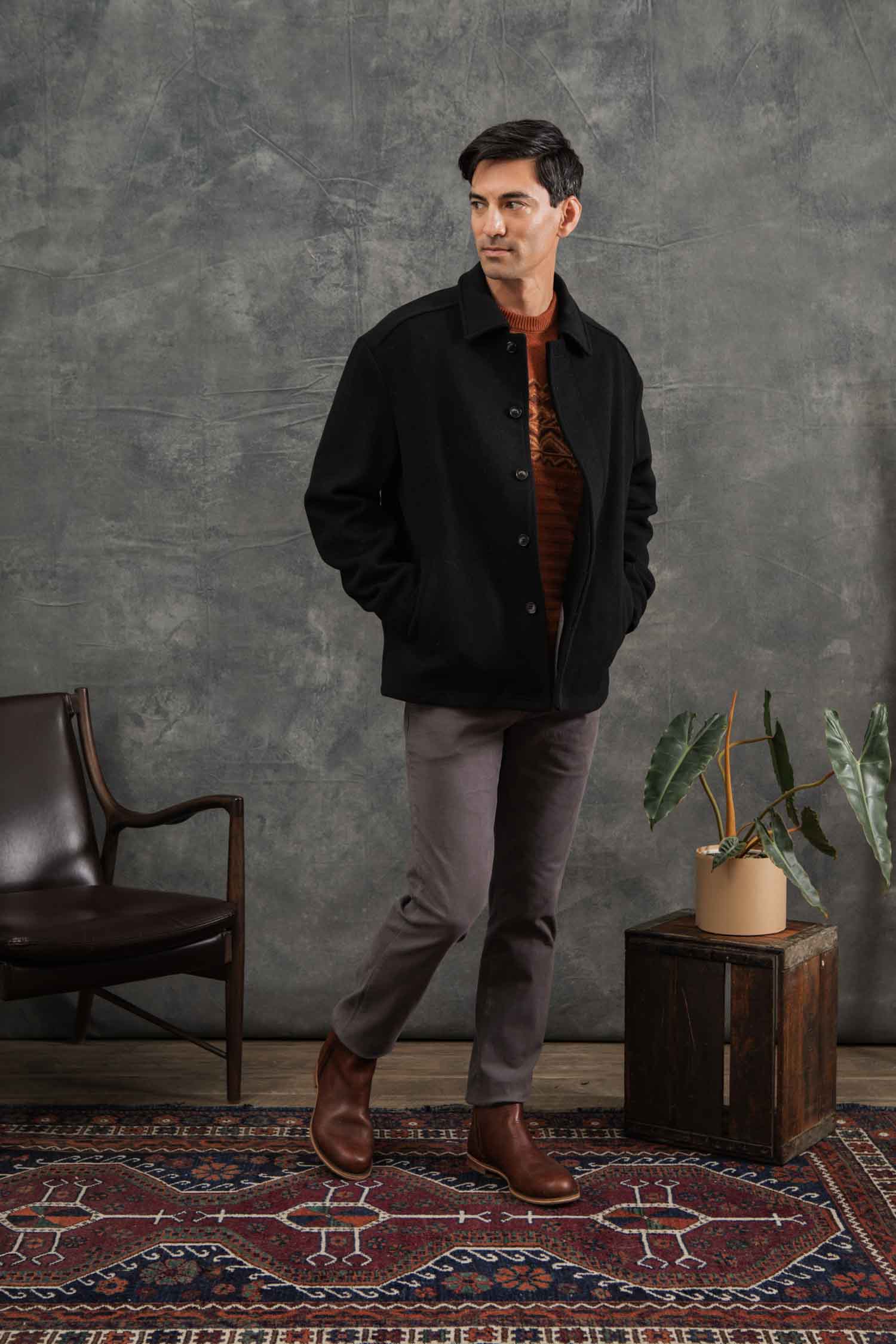 A man enjoys indoor comfort on a patterned rug, wearing the Bridge & Burn Clive Car Coat in black with his hands in his pockets, standing near a potted plant on a wooden box and a dark chair.