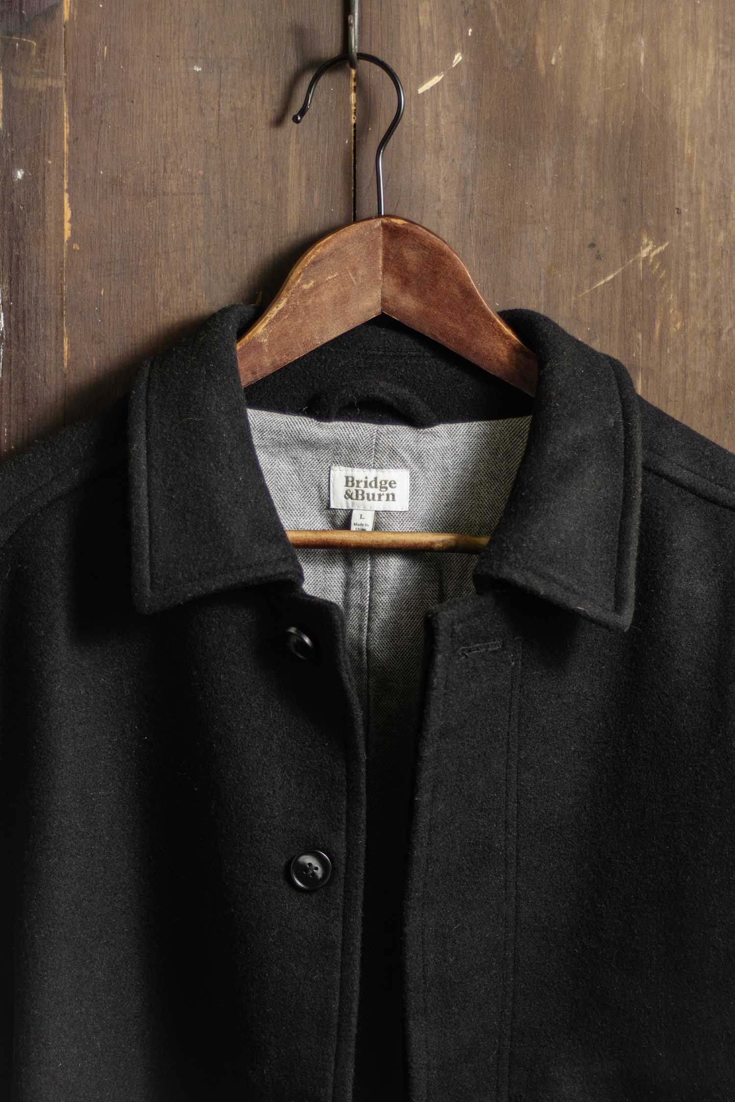 The Clive Car Coat in Black by Bridge & Burn hangs on a wooden hanger against a rustic wall, showcasing its heavyweight wool, collar design, black buttons, and visible label—perfect for timeless cold-weather style.