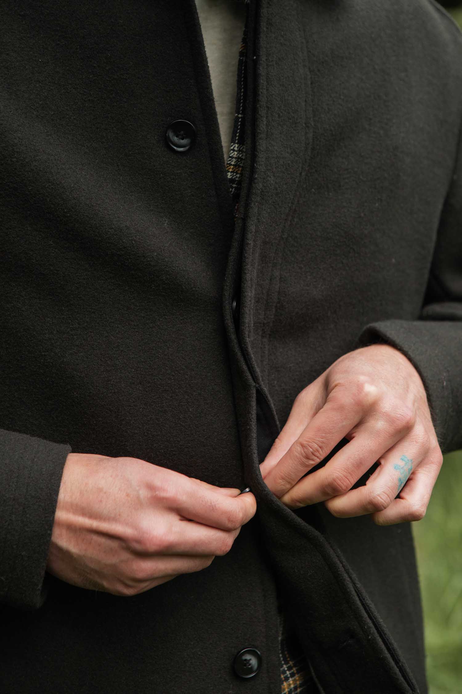 Only hands and torso are visible as someone buttons the Bridge & Burn Clive Car Coat in Deep Sage, revealing a small blue finger tattoo. The blurred green backdrop adds a cozy, cold-weather vibe.