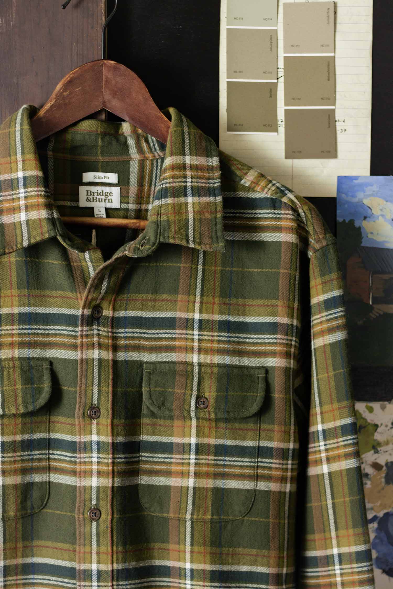 The Bridge & Burn Cole Slim Shirt in Green Mountain Plaid hangs on a wooden hanger, set against paint swatches, a rustic dark surface, and a partial painting of a building.