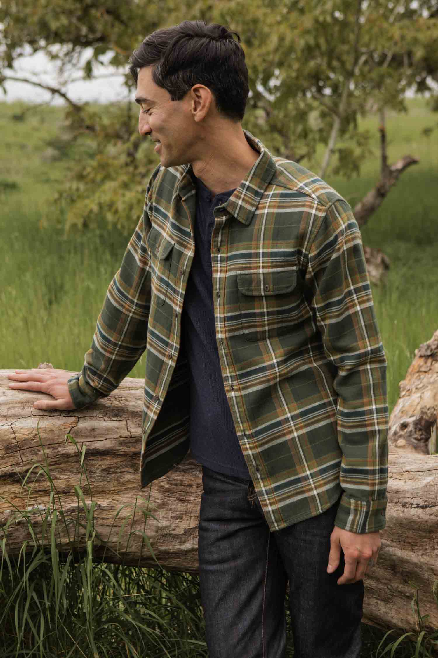 A man wearing the Bridge & Burn Cole Slim Shirt in Green Mountain Plaid stands outdoors, leaning on a fallen log and smiling with tall grass and a tree in the background.