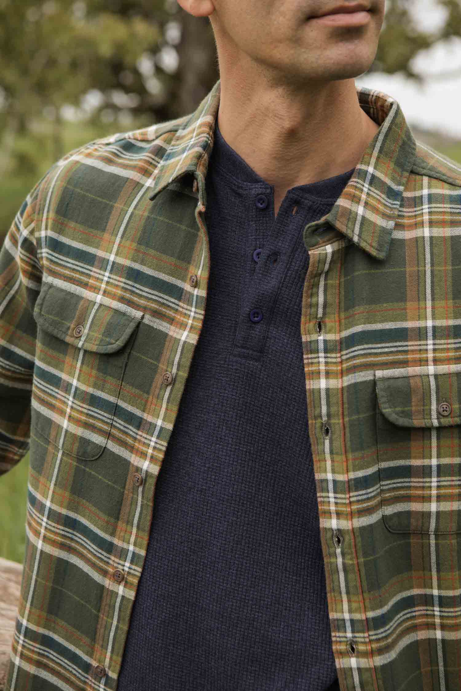 A person outdoors wears the Bridge & Burn Cole Slim Shirt in Green Mountain Plaid over a dark blue henley. The cropped image shows the torso and lower face with a blurred natural background.