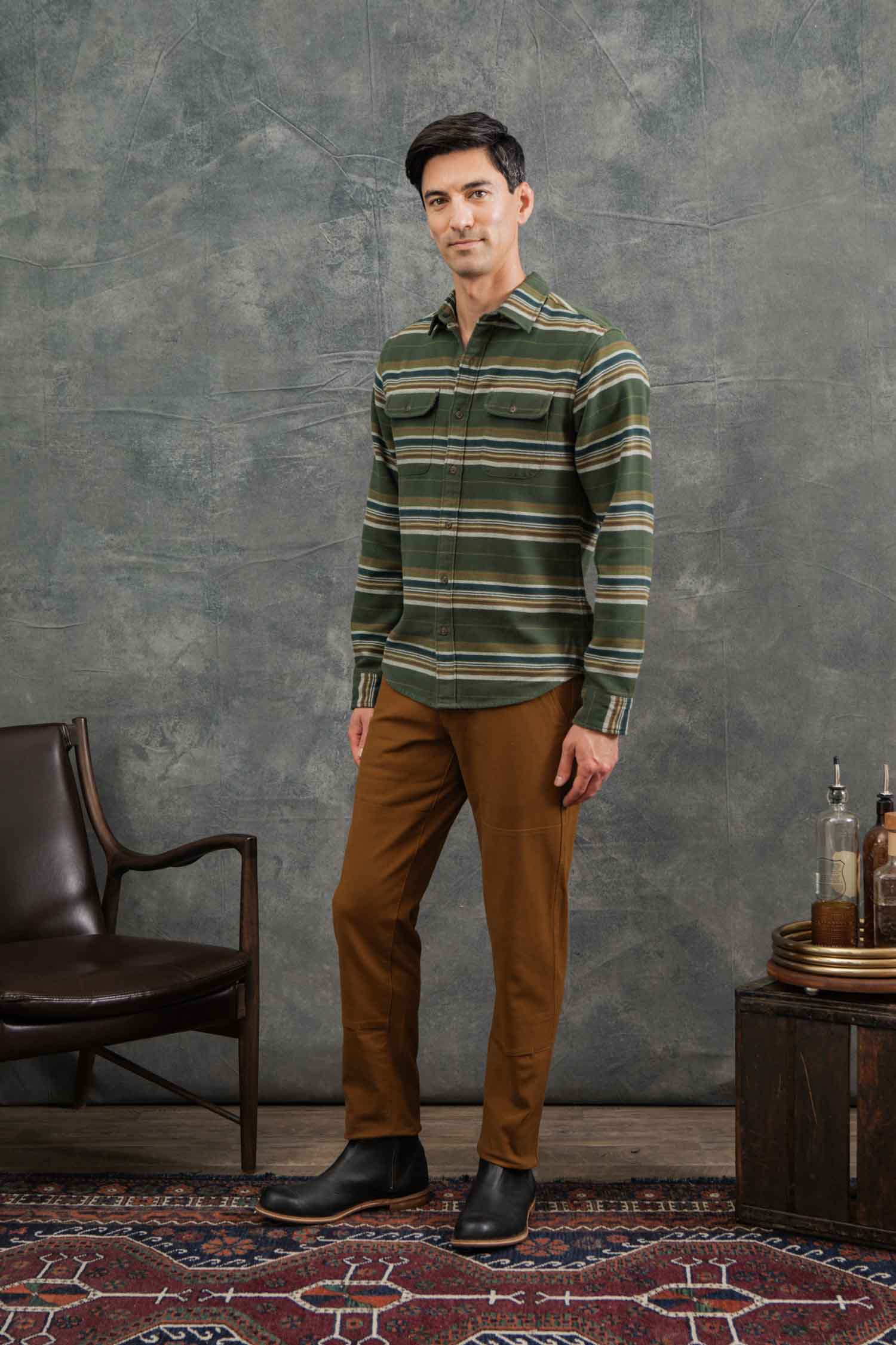 A man stands indoors on a patterned rug, wearing Bridge & Burn's Cole Slim Shirt in Wreath Stripe and brown pants, with black boots. He is beside a dark chair and small table with bottles, set against a textured gray backdrop.