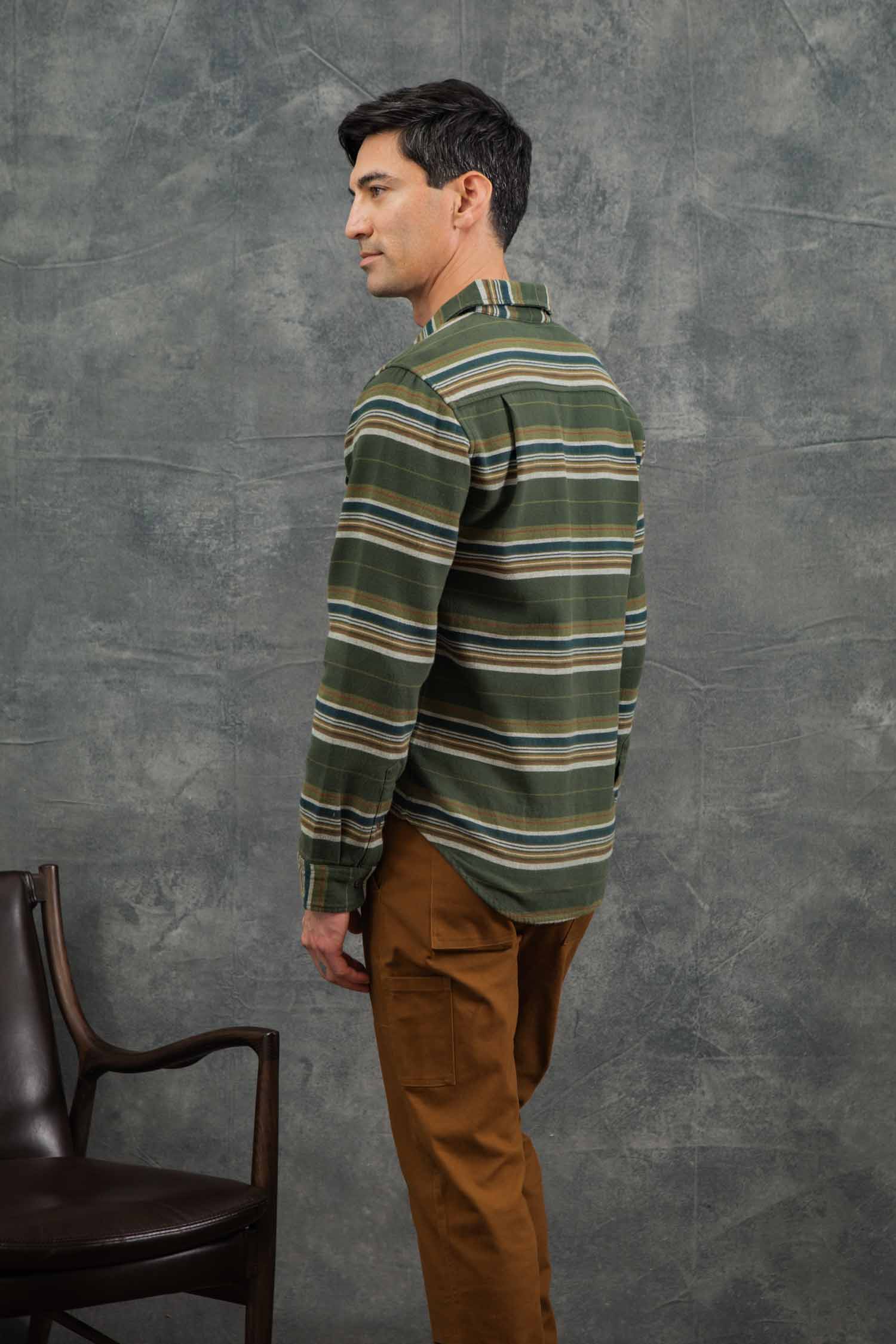 A man with short dark hair stands sideways, facing left. He wears the Bridge & Burn Cole Slim Shirt in Wreath Stripe and brown pants. A dark chair is to his left against a textured gray background.