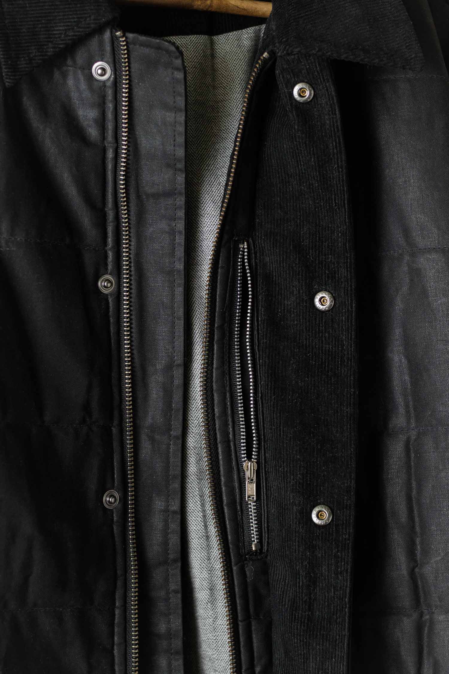 Close-up of the Bridge & Burn Holman Quilted Jacket in black waxed canvas, featuring metal snap buttons, a partially unzipped front showing the light gray lining, and a textured outer finish.