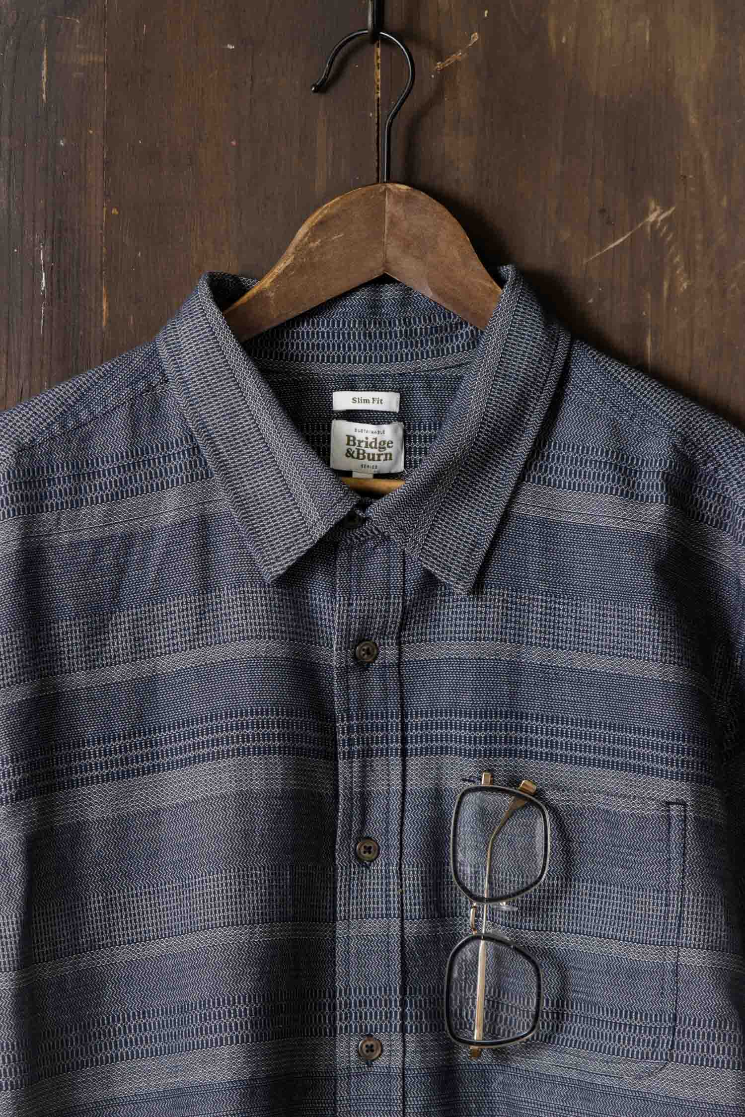 The Bridge & Burn Jude Shirt in Cascade Weave, a classic fit men's button-up with a blue pattern, hangs on a wooden hanger against a dark wood background. Eyeglasses are hooked onto the shirt’s front pocket.