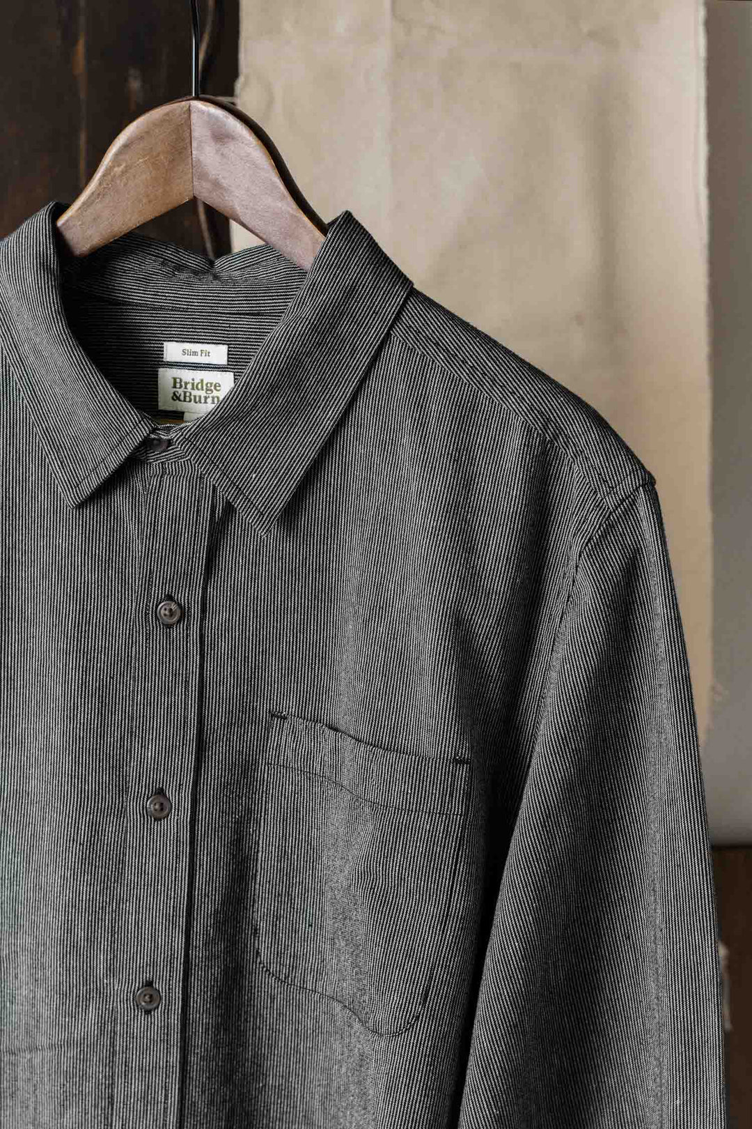 A close-up of the Bridge & Burn Jude Shirt in Onyx Stripe—classic fit, 100% cotton with thin vertical stripes—hangs on a wooden hanger, showcasing its collar and chest pocket against a softly blurred, neutral background.