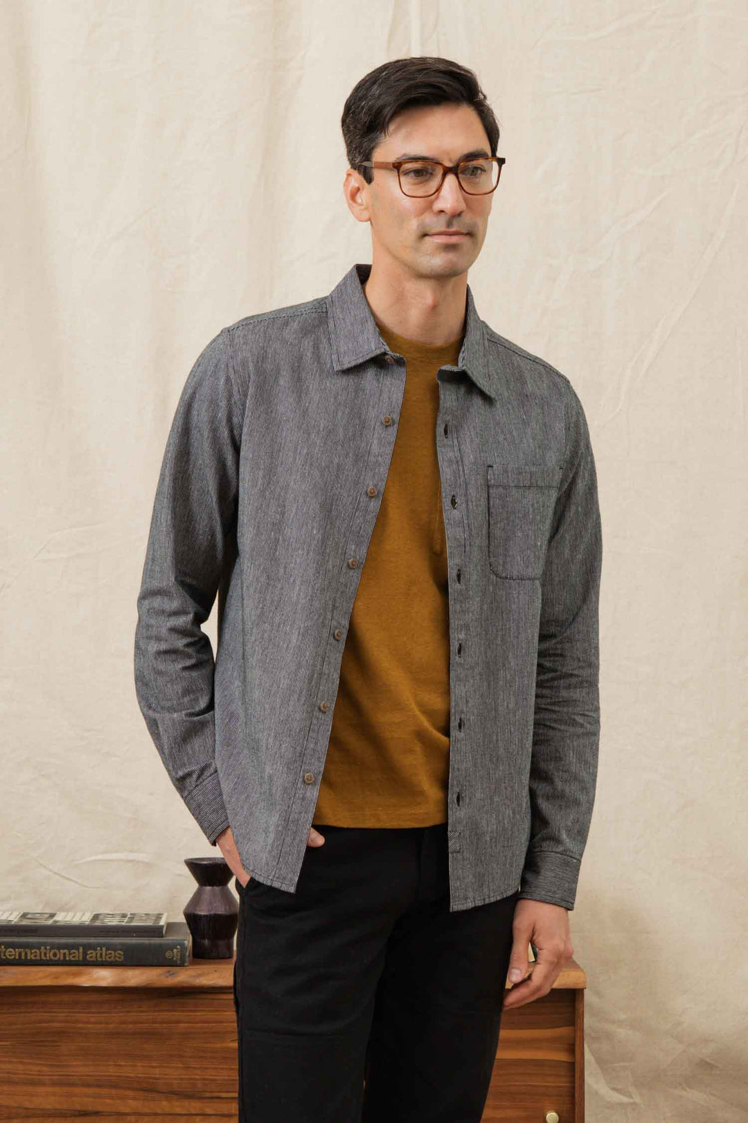 A man with short dark hair and glasses stands indoors wearing the Bridge & Burn Jude Shirt in Onyx Stripe over a brown tee and black pants, posed before a beige backdrop beside a wooden table with books and decor.