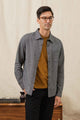 A man with short dark hair and glasses stands indoors wearing the Bridge & Burn Jude Shirt in Onyx Stripe over a brown tee and black pants, posed before a beige backdrop beside a wooden table with books and decor.