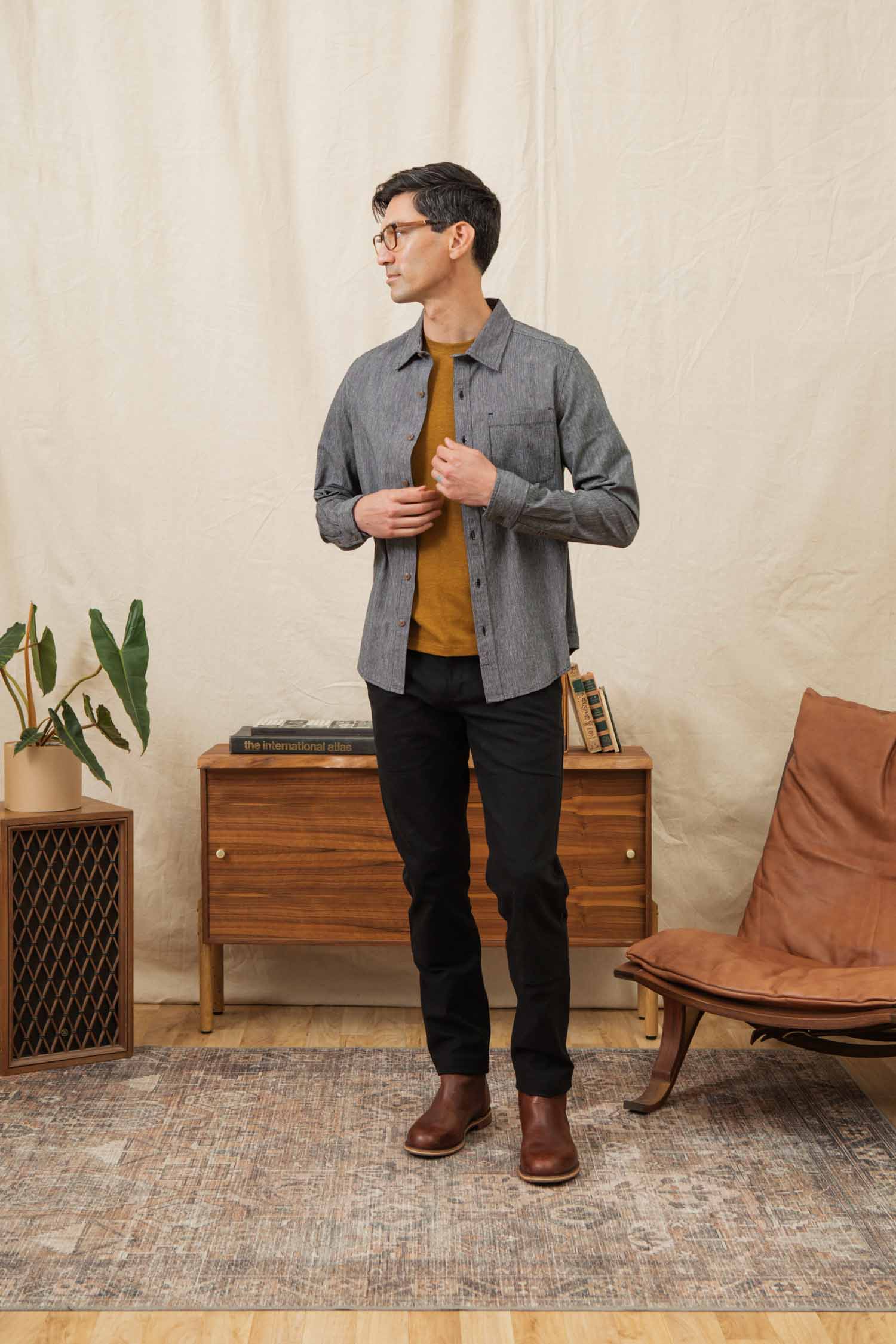 A man wearing glasses, the Bridge & Burn Jude Shirt in Onyx Stripe over a mustard tee, black pants, and brown boots stands in a stylish living space with a wooden cabinet, plant, brown chair, and beige backdrop.