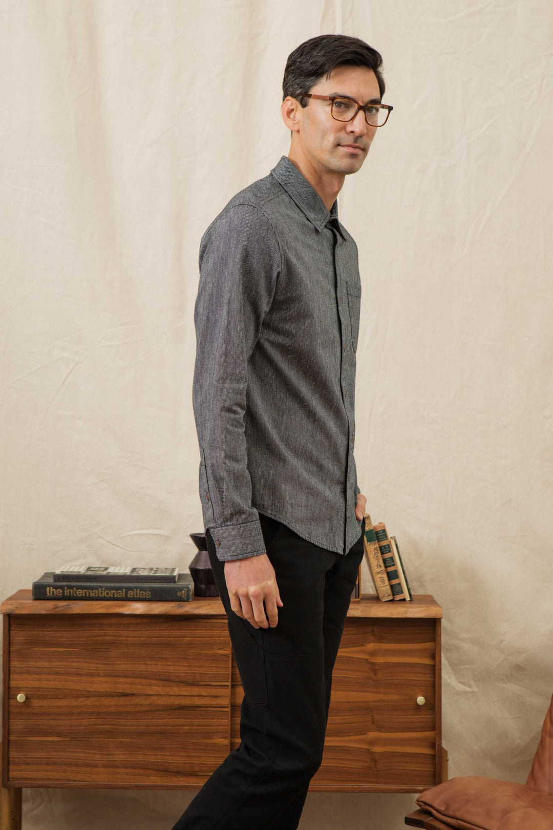 A man with short dark hair and glasses stands in profile, wearing the Bridge & Burn Jude Shirt in Onyx Stripe and black pants. He is beside a mid-century wooden cabinet topped with books and small decorative objects.