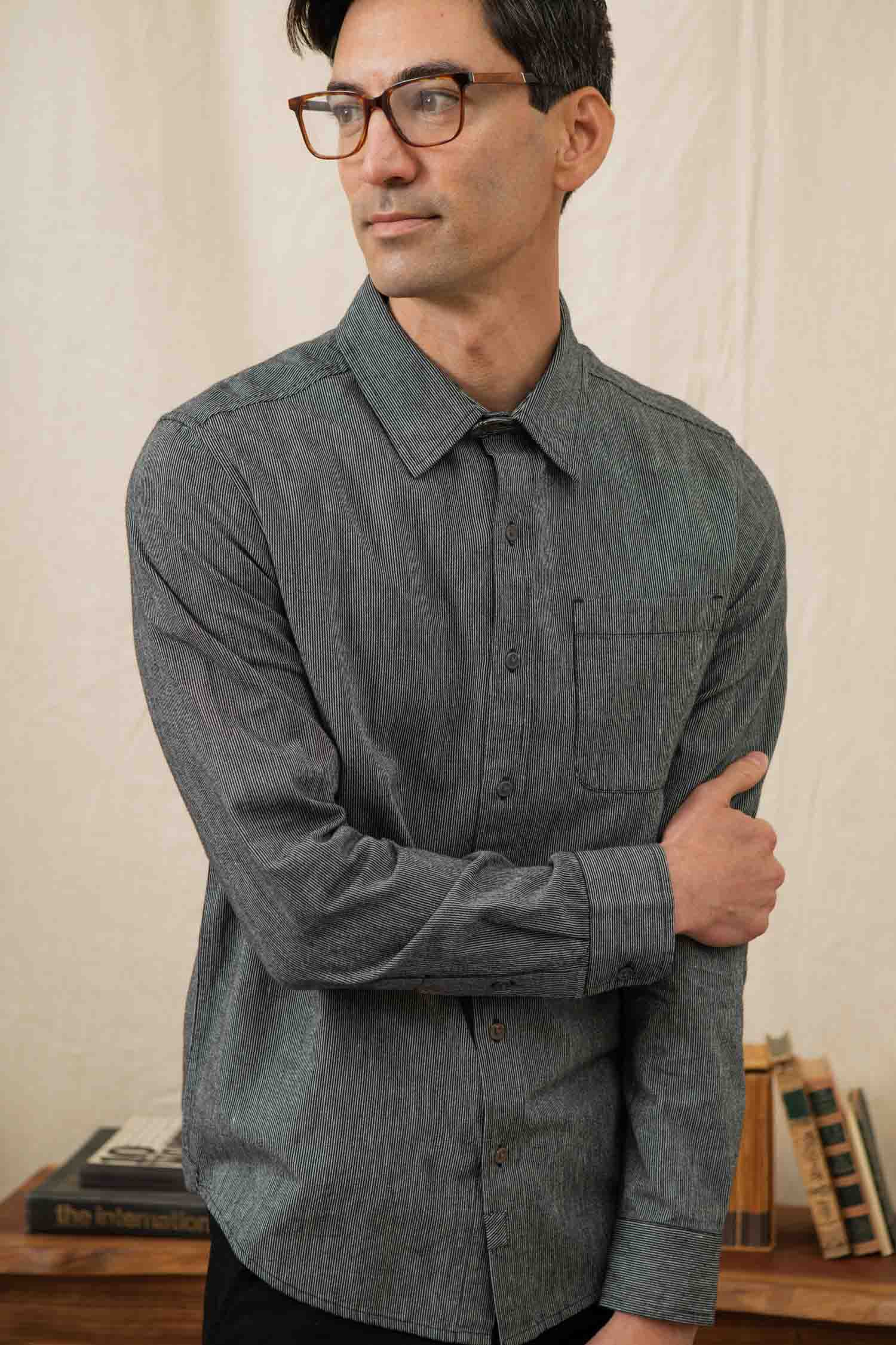 A man with short dark hair and glasses wears the Bridge & Burn Jude Shirt in Onyx Stripe, a classic fit men's button-up with a chest pocket. He stands indoors, looking to the side, holding his arm, with books stacked behind him.