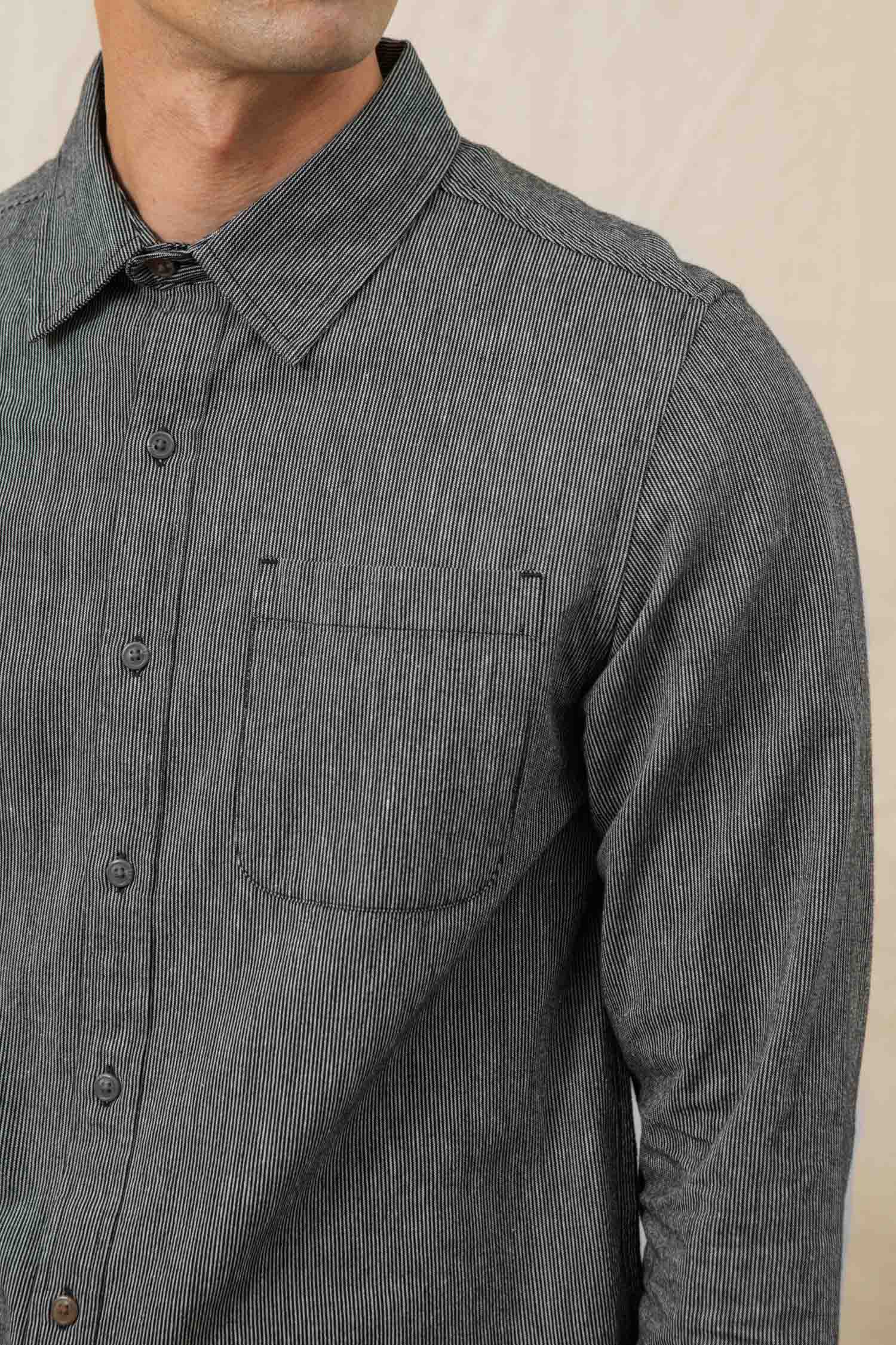 A close-up shows a person wearing the Bridge & Burn Jude Shirt in Onyx Stripe—men's, gray pinstripe, 100% cotton, with long sleeves and a small chest pocket. Only the torso and lower face are visible.