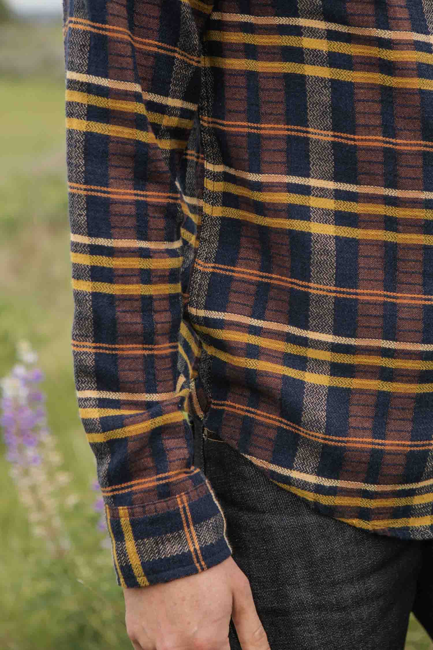 A person in the Bridge & Burn Larson Shirt / Desert Dawn Plaid, a classic fit long-sleeve flannel in brown, yellow, and navy, stands outdoors on grass with their arm relaxed by their side. Dark pants and blurred flowers are partially visible.