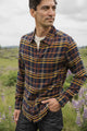A man wearing the Bridge & Burn Larson Shirt in Desert Dawn Plaid and jeans stands in a grassy field with purple wildflowers, looking to the side. The sky is cloudy and the background is softly blurred.