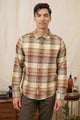 A man with dark hair wears the Bridge & Burn Larson Shirt in Latte Plaid and brown pants, standing indoors in front of a light curtain with wooden furniture and decor behind him.