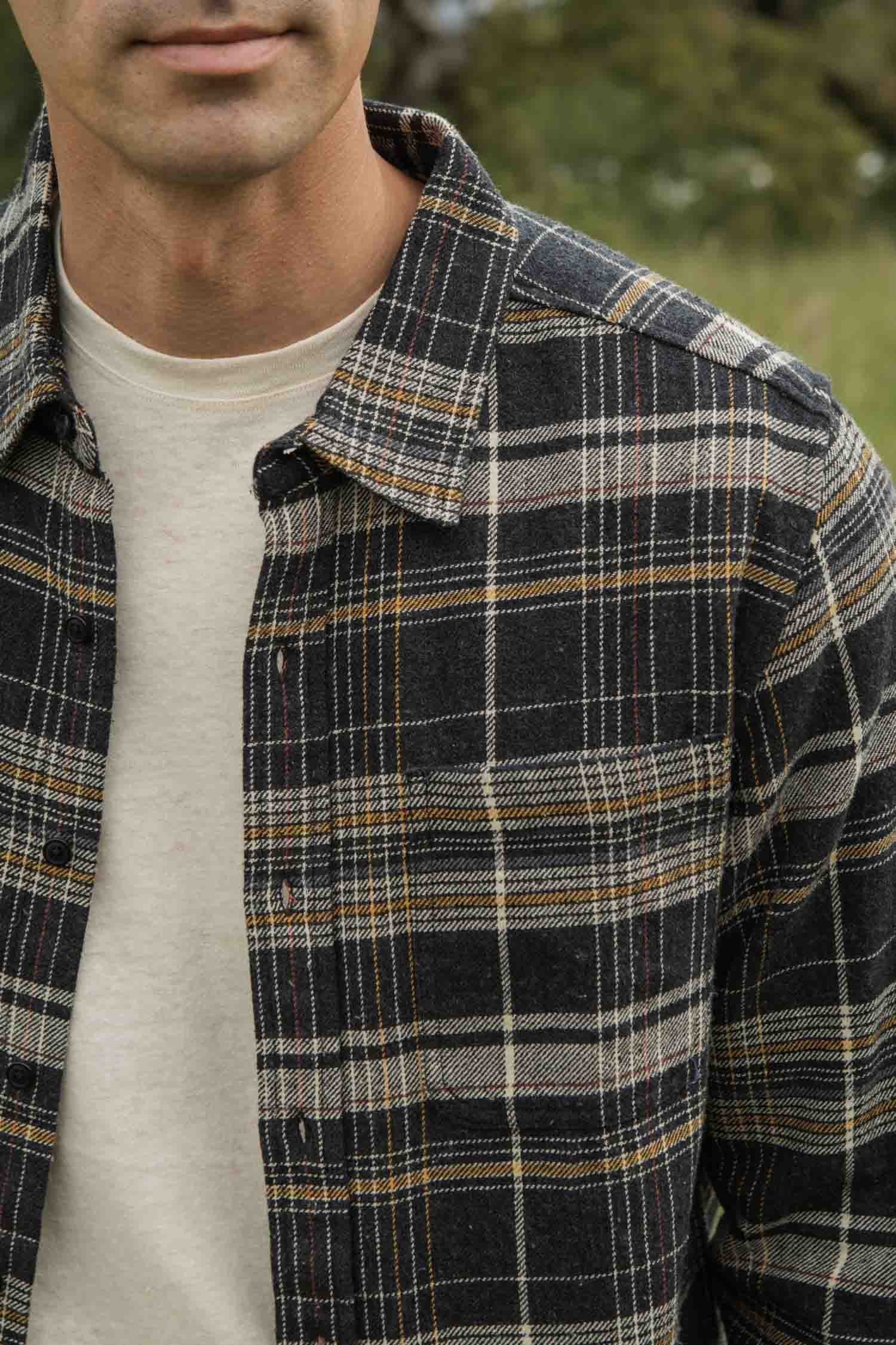 A person stands outdoors, wearing the Bridge & Burn Larson Shirt in Volcanic Plaid over a cream T-shirt; only the lower face and upper torso are visible, with blurred greenery in the background.