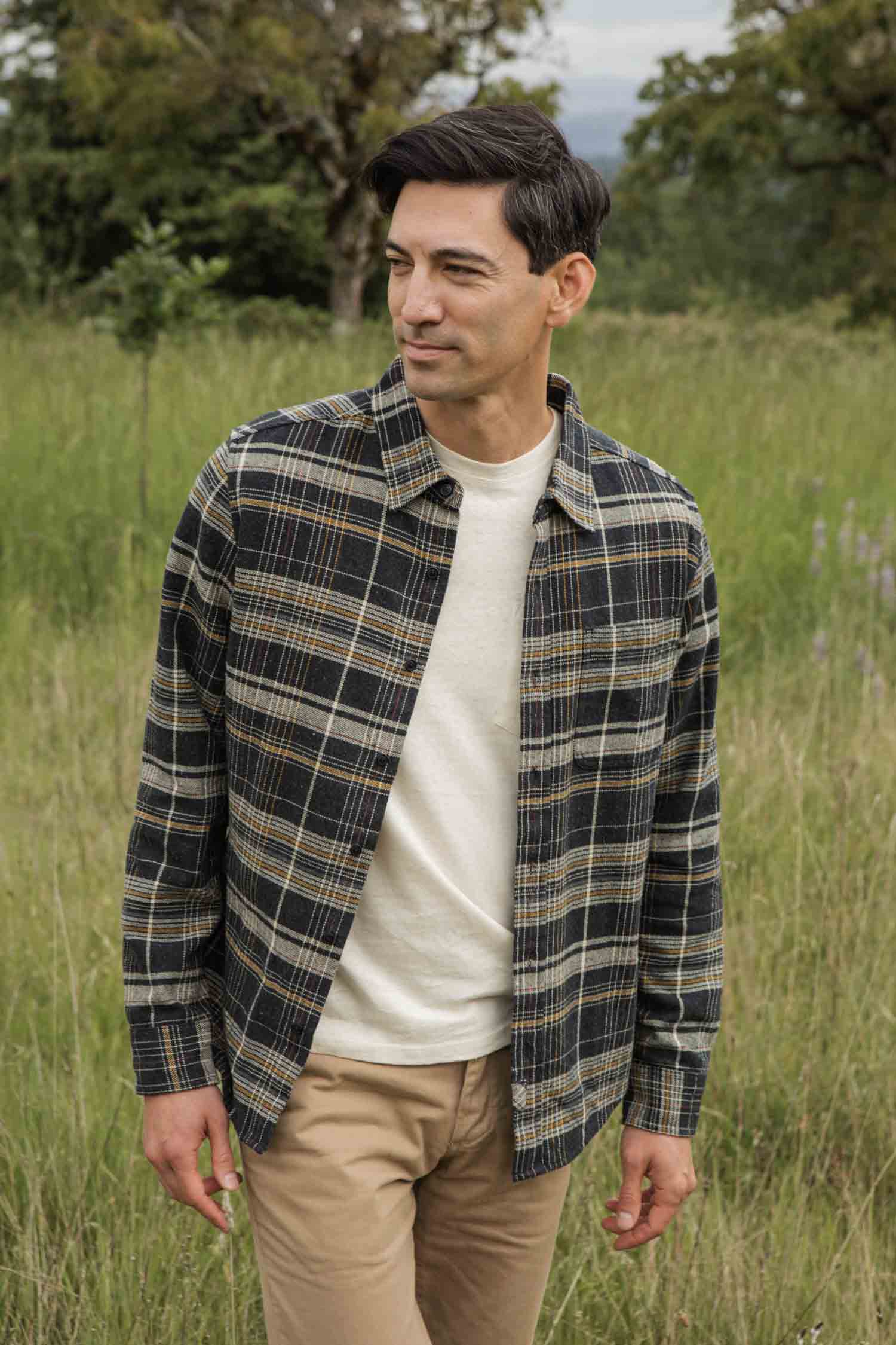 A man stands in a grassy field, smiling slightly as he looks left. He wears the Bridge & Burn Larson Shirt in Volcanic Plaid over a cream t-shirt and beige pants. Trees and greenery are visible in the background.