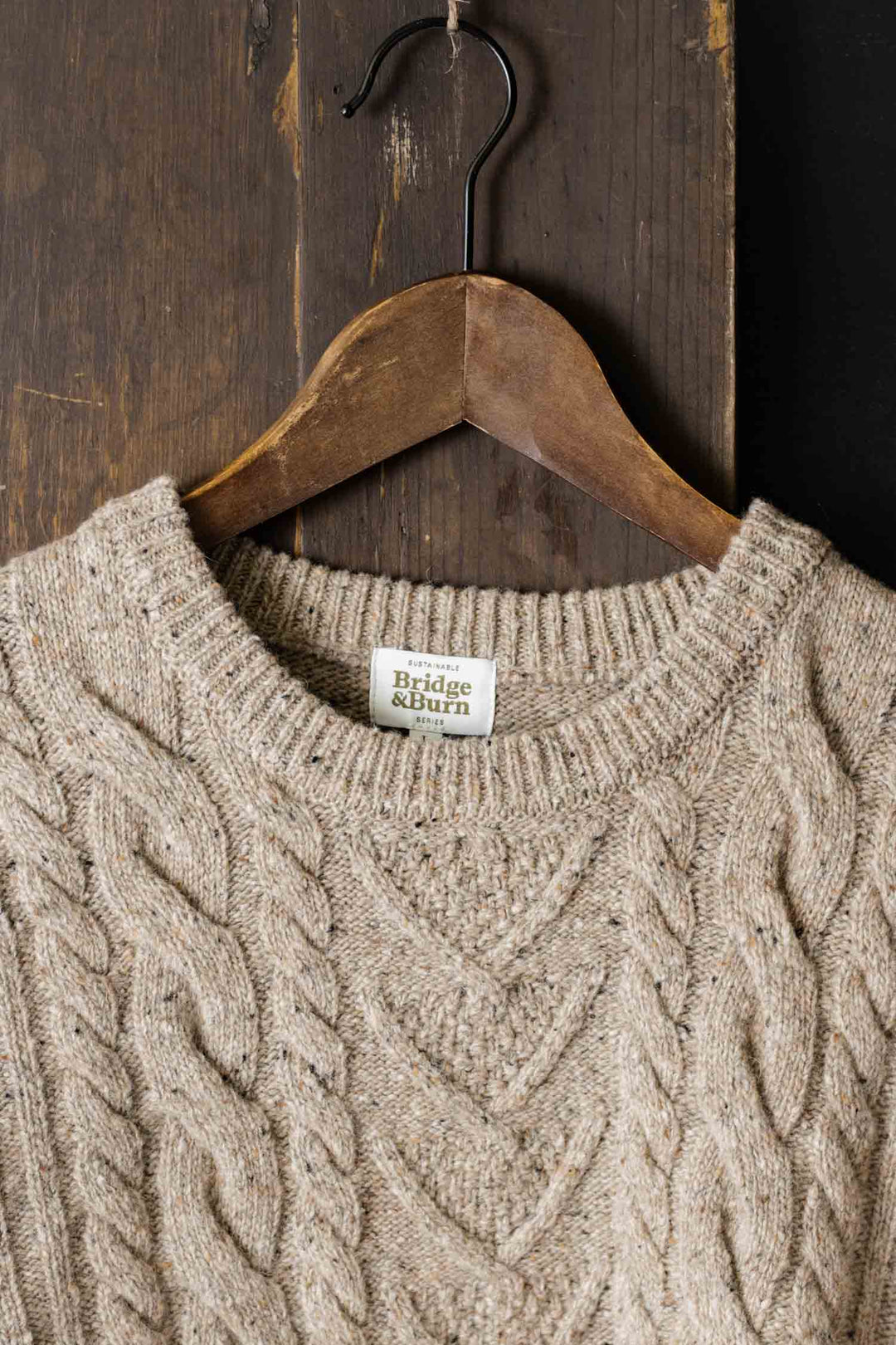 The Ludlow Sweater in Flecked Oatmeal by Bridge & Burn, crafted from RWS-certified wool, hangs on a wooden hanger against a rustic wooden background.