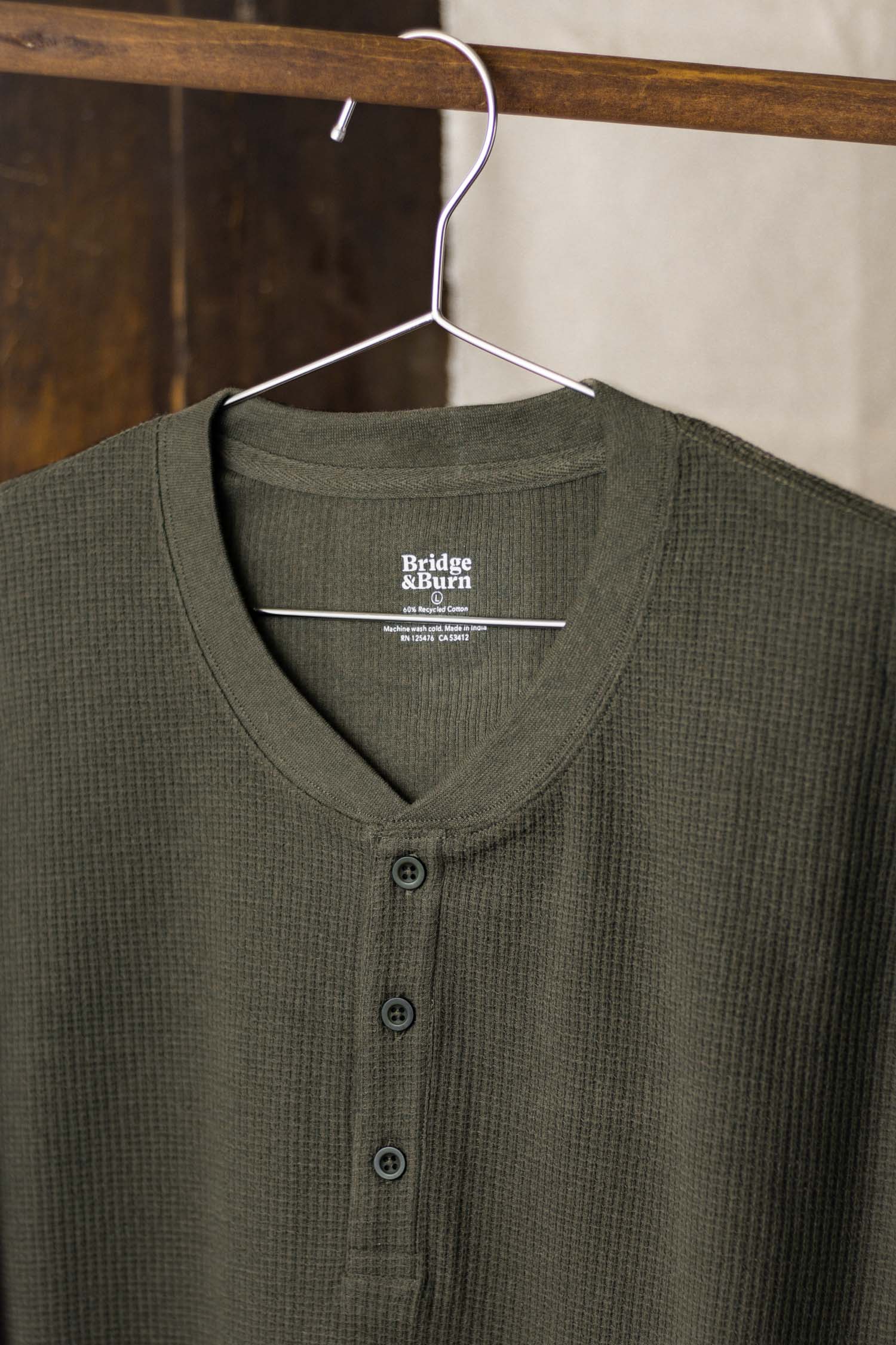 The Bridge & Burn Men's Recycled Waffle Henley in Forest Melange, featuring three buttons and a waffle-knit texture, hangs on a metal hanger against a neutral indoor background.