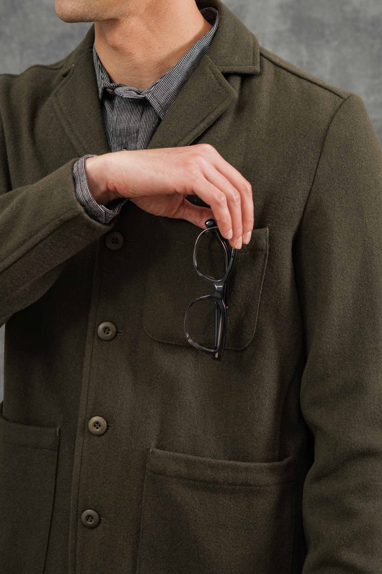 A man in a Bridge & Burn Nate Chore Blazer in Basque Green Wool wears a striped shirt and holds black eyeglasses by his chest pocket, his face partially out of frame against a gray background.