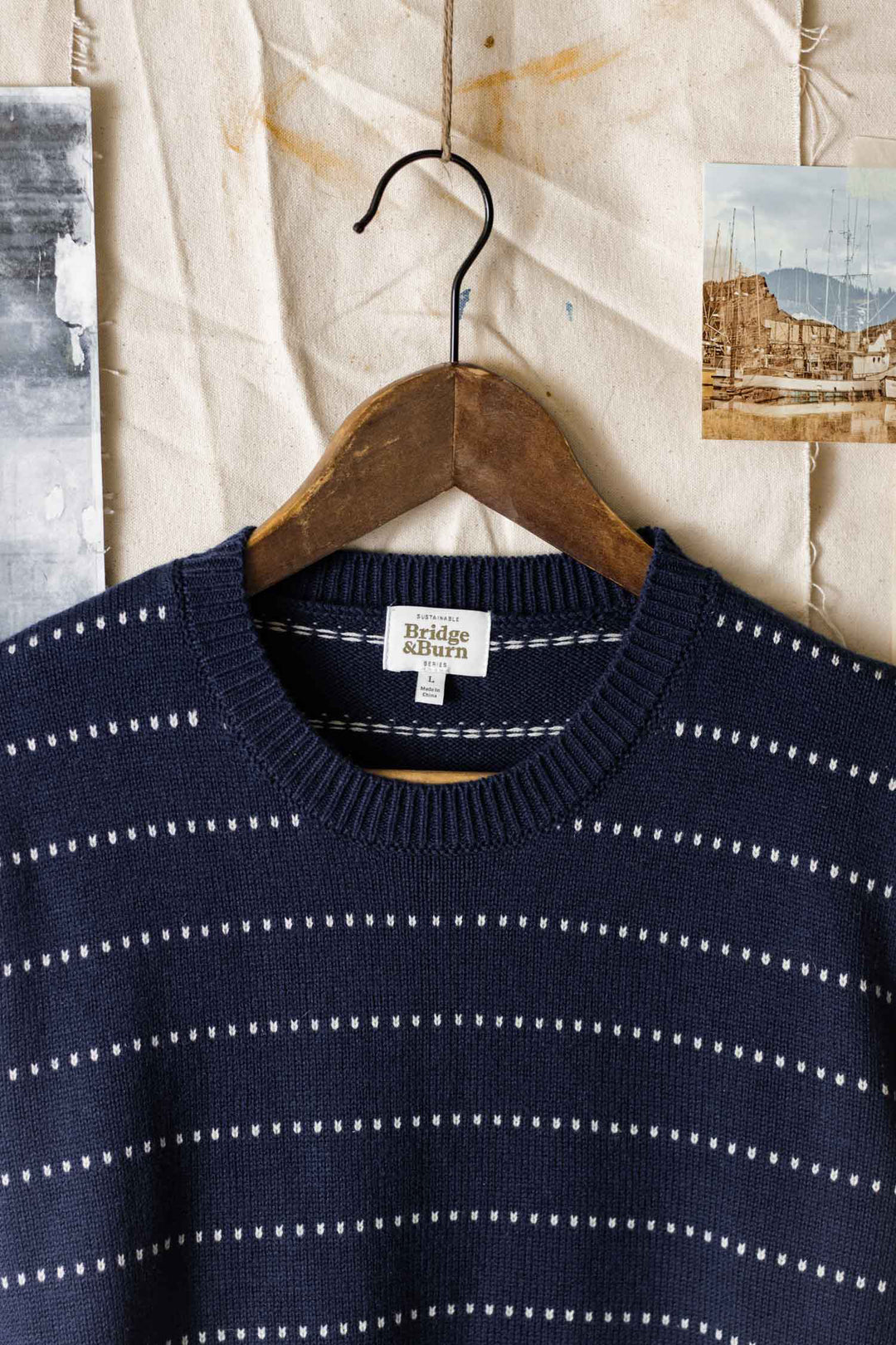 The Orson Sweater in Navy Dotted Path by Bridge & Burn hangs on a wooden hanger against a beige textured background, flanked by two vintage-style photographs.