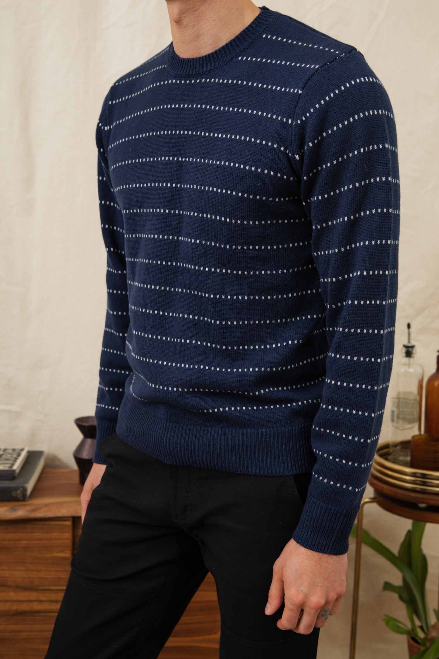 A person stands indoors near a wooden table with decor, wearing Bridge & Burn's Orson Sweater in Navy Dotted Path and black pants.