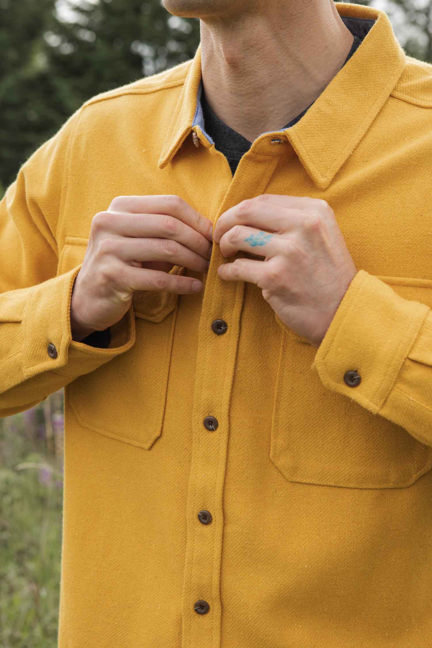 A person fastens the buttons of the Bridge & Burn Pointer Overshirt in Antique Gold, made from recycled fibers. Only their upper body is visible against a backdrop of blurred greenery.