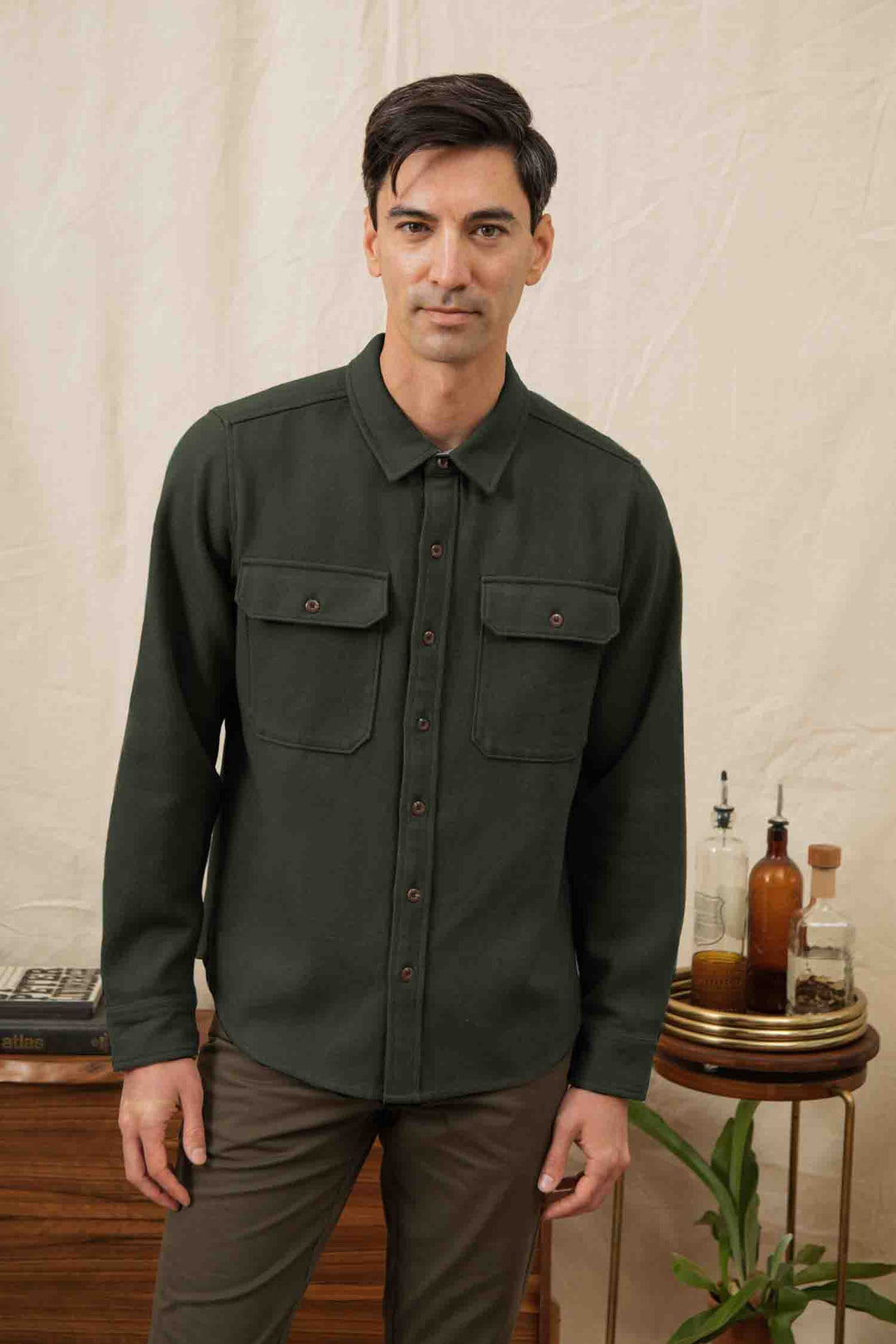 A man with dark hair stands indoors in front of a beige backdrop, wearing the Bridge & Burn Pointer Overshirt in Dark Sage and brown pants. Behind him, bottles, a small plant, and books sit on a wooden table.