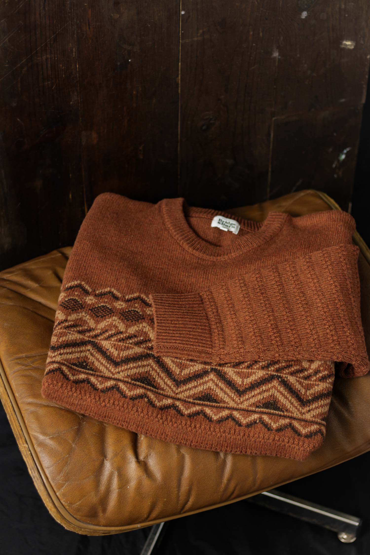 The Rockwood Fairisle Sweater in Walnut Grove by Bridge & Burn, featuring a geometric zigzag pattern in earthy tones, is folded on a worn tan leather chair set against a dark wooden backdrop.