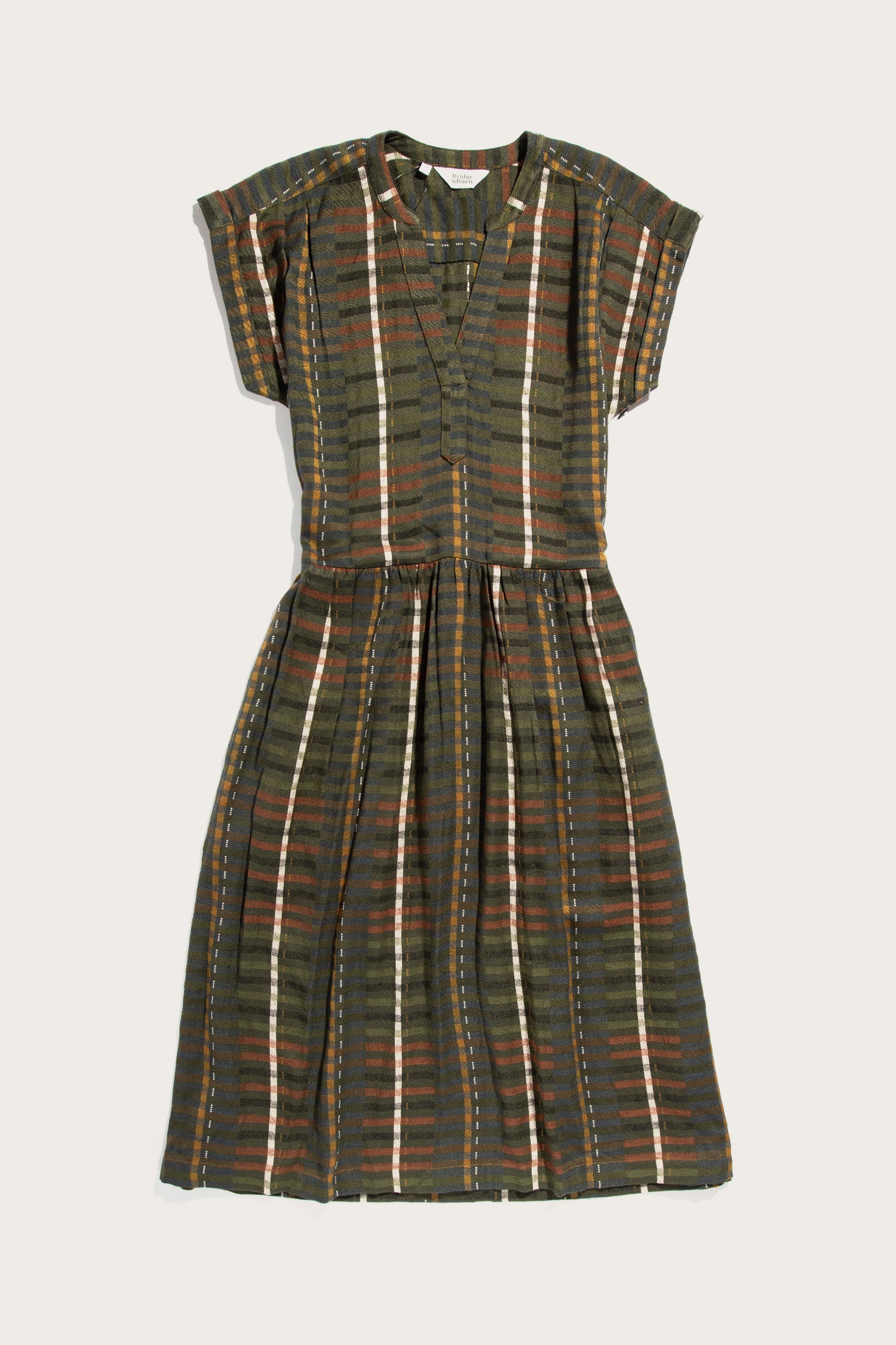 The Bridge & Burn Ira Dress in Olive Stripe is a short-sleeve, knee-length pullover with a V-neck and fitted waist, made from a modal-linen blend featuring dark green, brown, white, and rust plaid stripes.