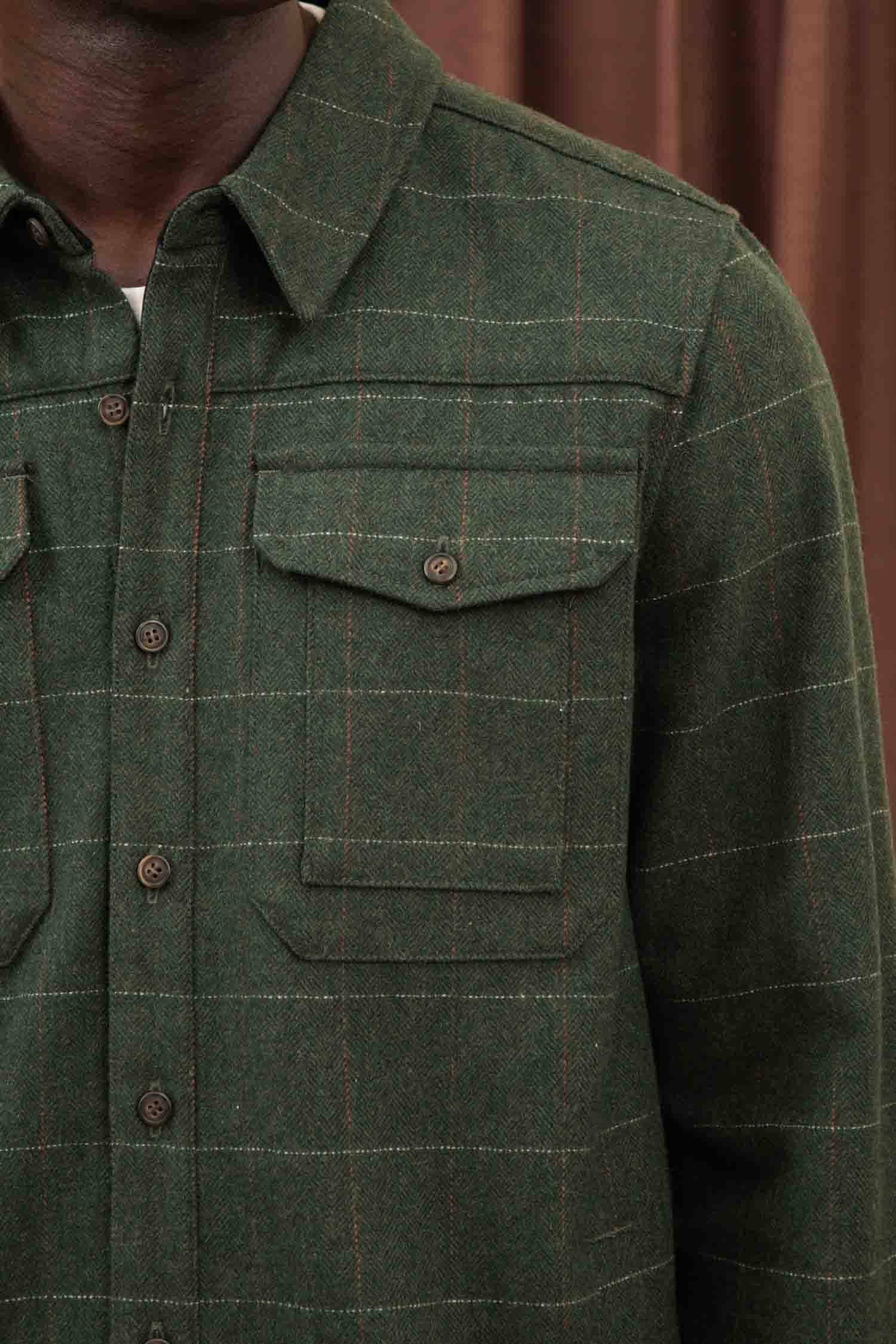 A close-up of a person wearing the Bridge & Burn Fielding Shirt Jacket in Cypress Windowpane—a green flannel with yellow and red grid lines and two chest pockets. Brown curtains are visible in the background.