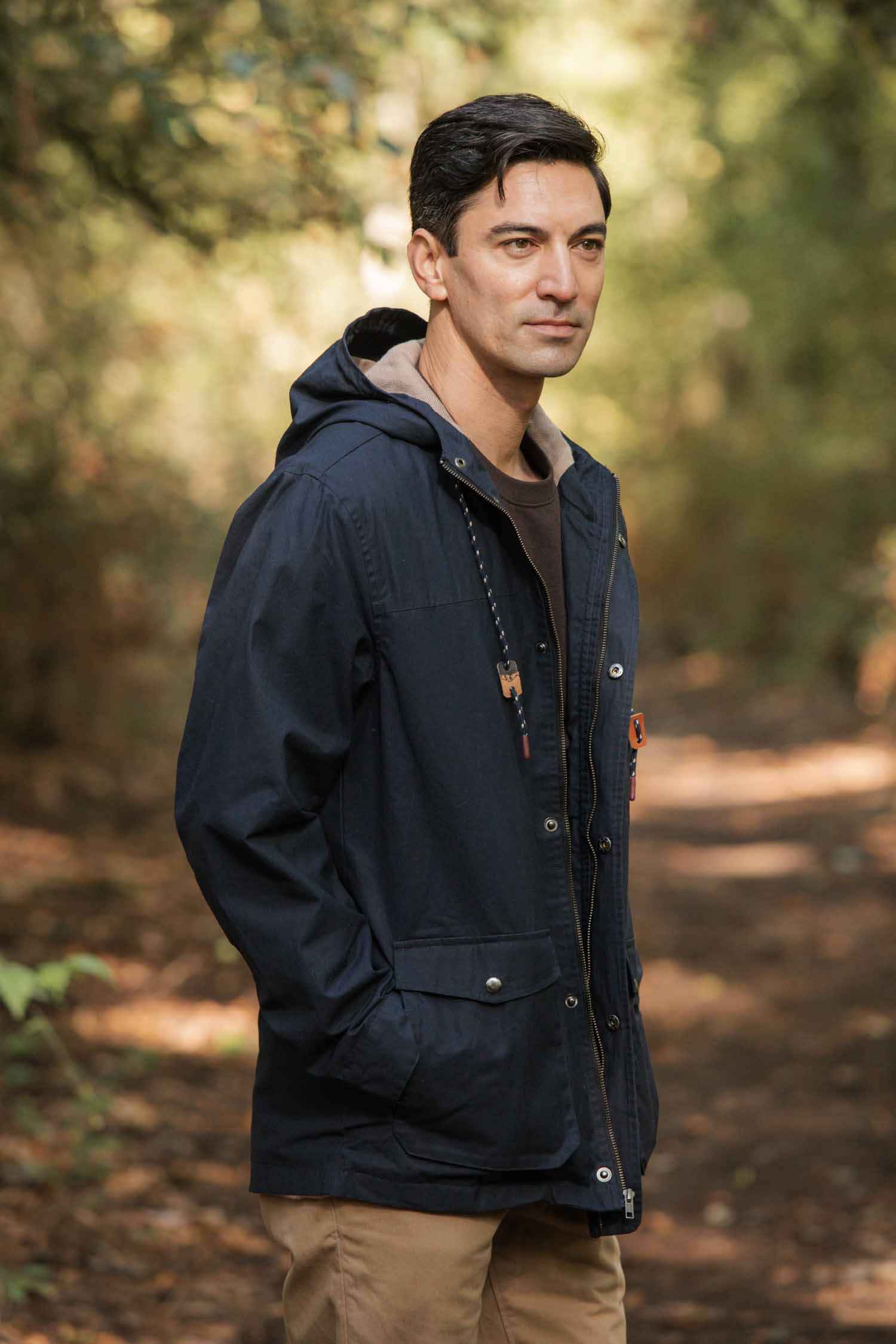 A man in a Bridge & Burn Marshall Jacket in navy waxed canvas and khaki pants stands on a forest path, surrounded by trees, with his hands in his pockets and a neutral expression.
