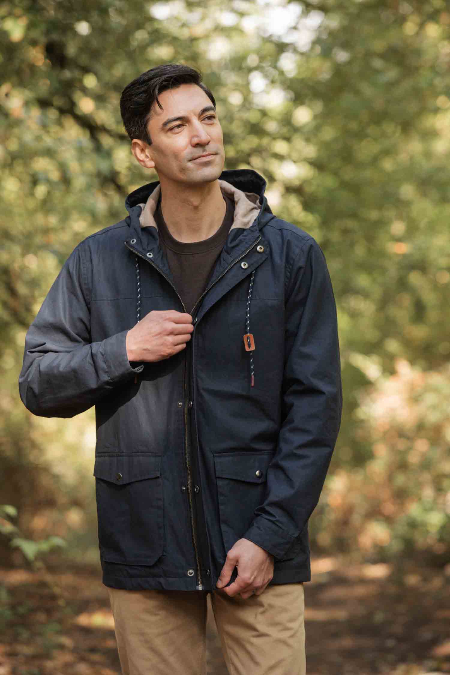 A man stands outdoors in a wooded area, smiling as he adjusts the zipper of his Bridge & Burn Marshall Jacket in navy waxed canvas—paired with tan pants for a classic, rugged look.