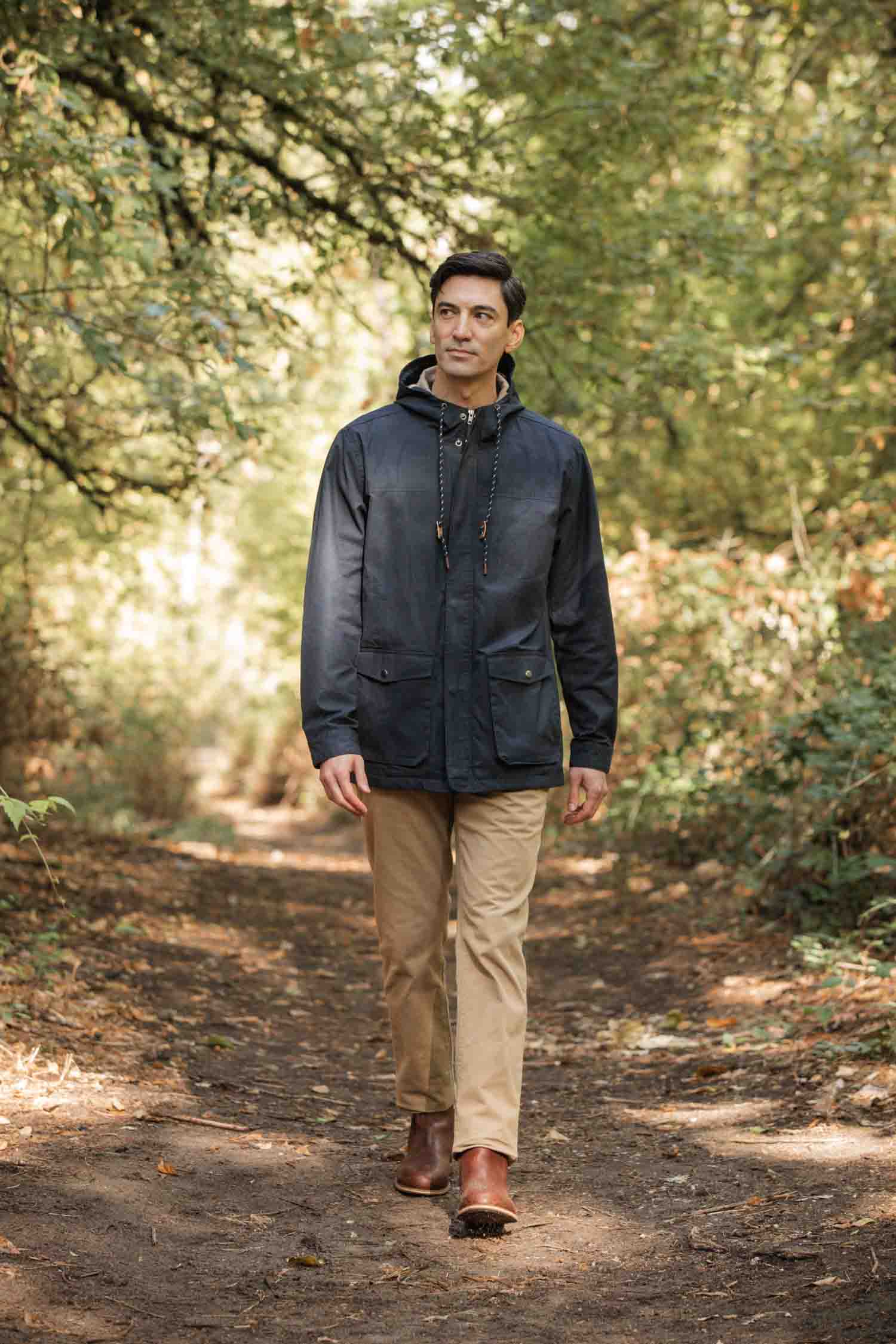 A man wearing a Bridge & Burn Marshall Jacket in navy waxed canvas, beige pants, and brown boots walks along a dirt path through a green, leafy forest on a sunny day.