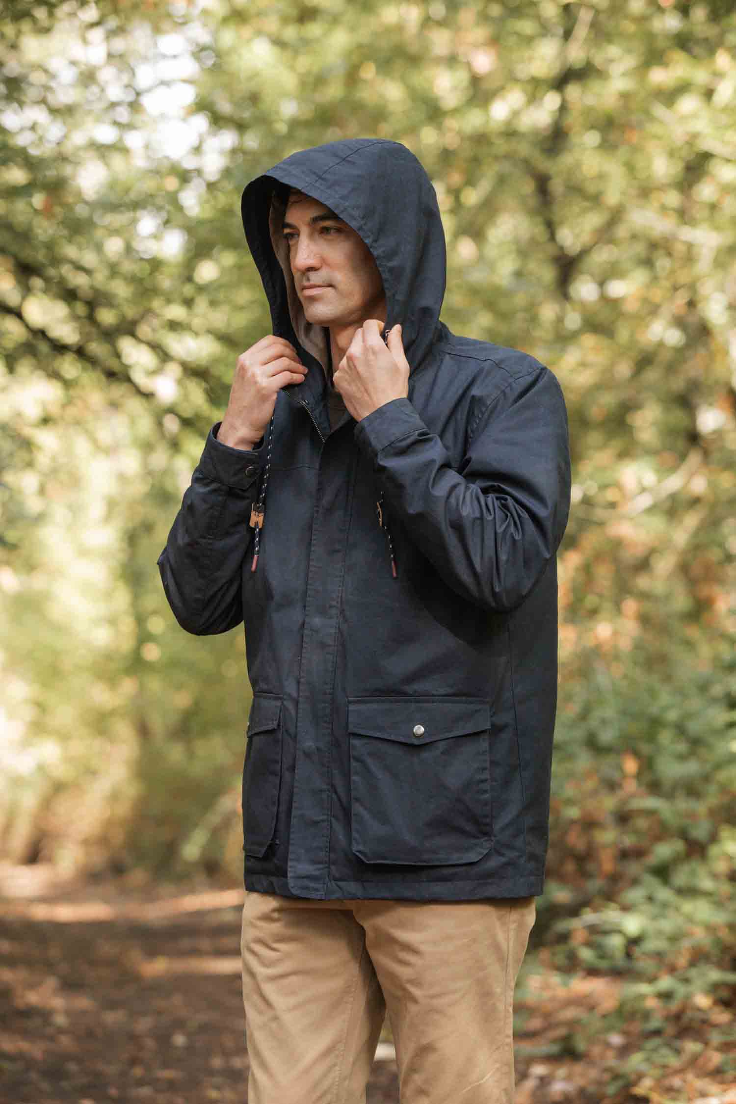 A man stands on a forest path in soft natural light, wearing the Bridge & Burn Marshall Jacket in navy waxed canvas and khaki pants, holding the sides of his hood and surrounded by lush greenery.