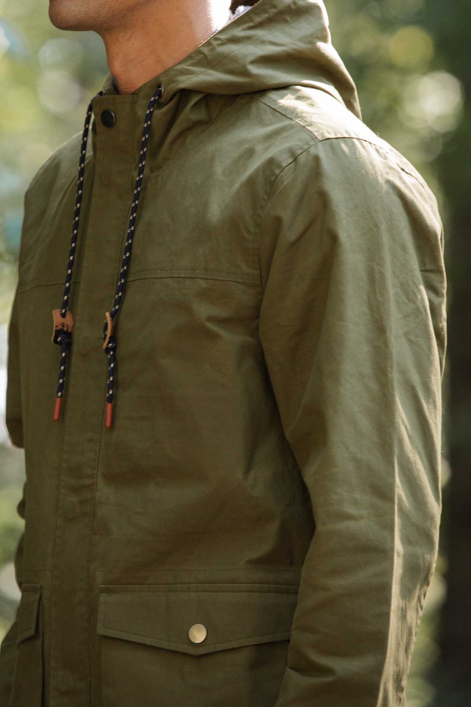 A person wearing the Bridge & Burn Marshall Jacket in olive waxed canvas, featuring black and white drawstrings and buttoned pockets, stands outdoors in soft natural light.
