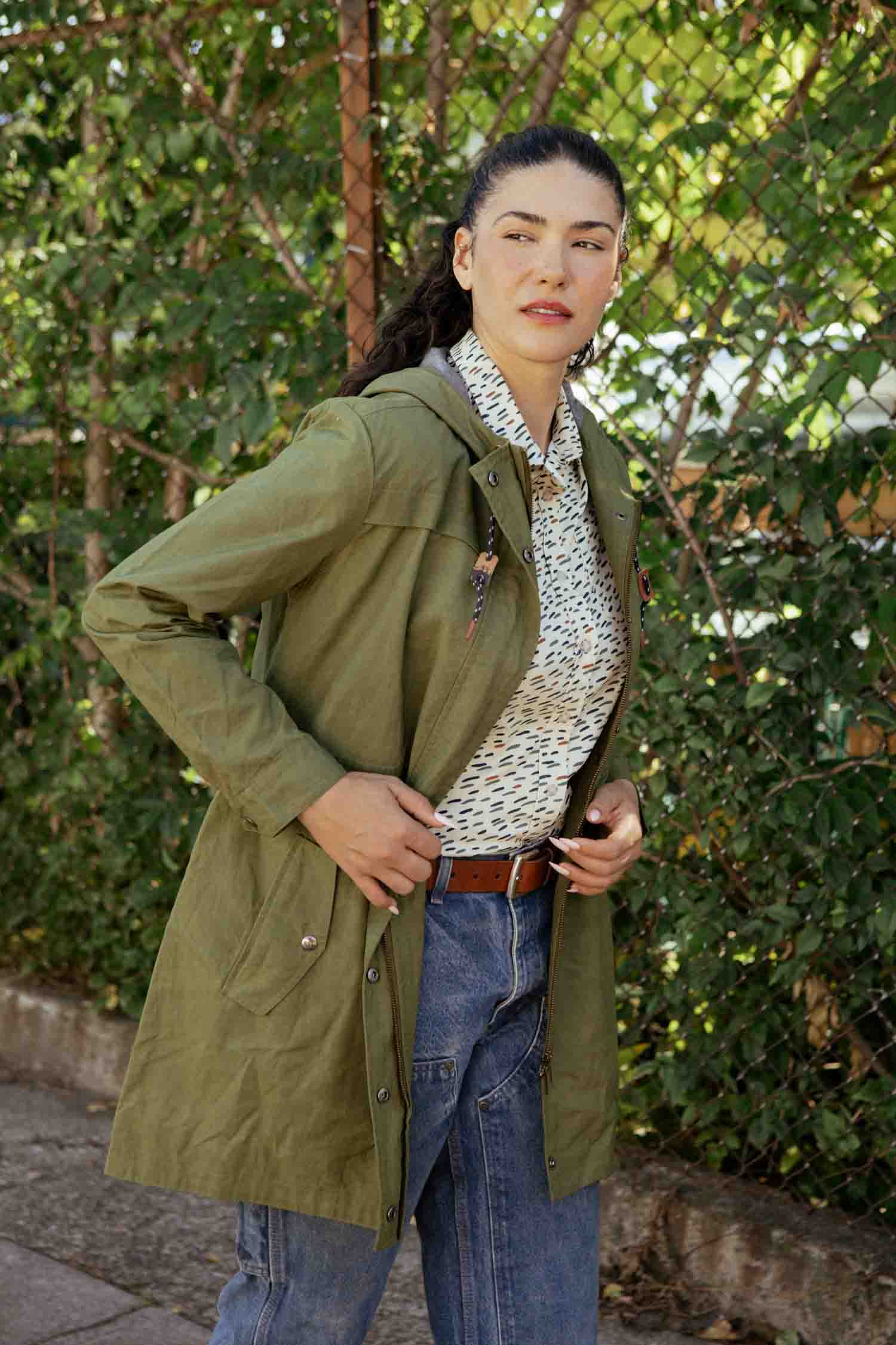 A woman stands outdoors in front of greenery, wearing the Bridge & Burn Briar Jacket in olive waxed canvas over a patterned shirt, blue jeans, and a brown belt. She looks to the side with one hand on her jacket.