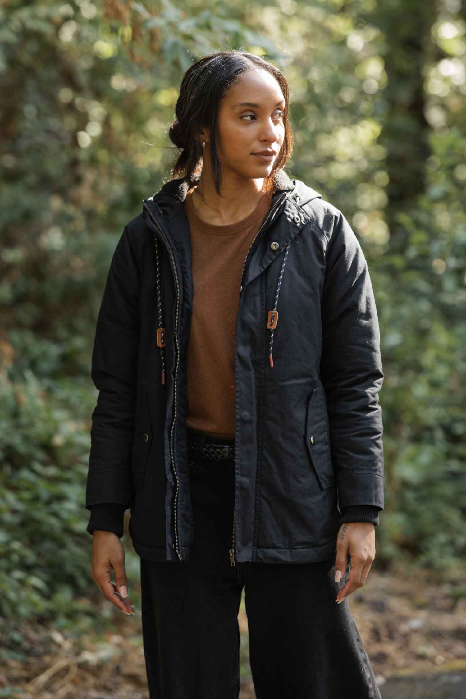 A woman with wavy hair stands outdoors in sunlight, wearing the Bridge & Burn Sequoia Parka in Black Waxed Canvas over a brown shirt, her hands relaxed at her sides as she looks slightly to the right among wooded trees.